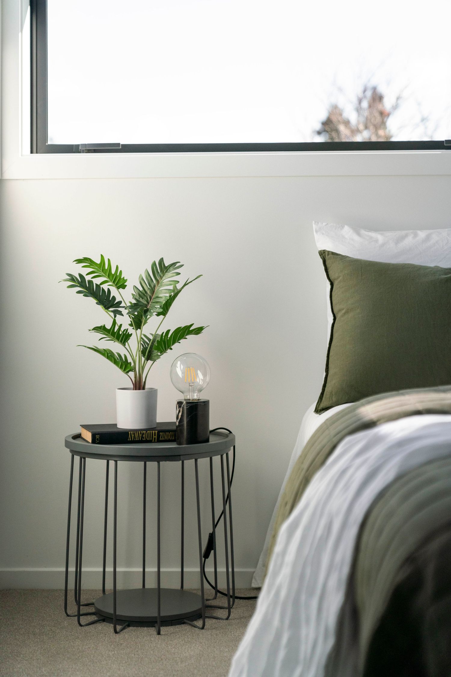 Minimalist bedside table with potted plant and globe lamp next to olive green pillow.
