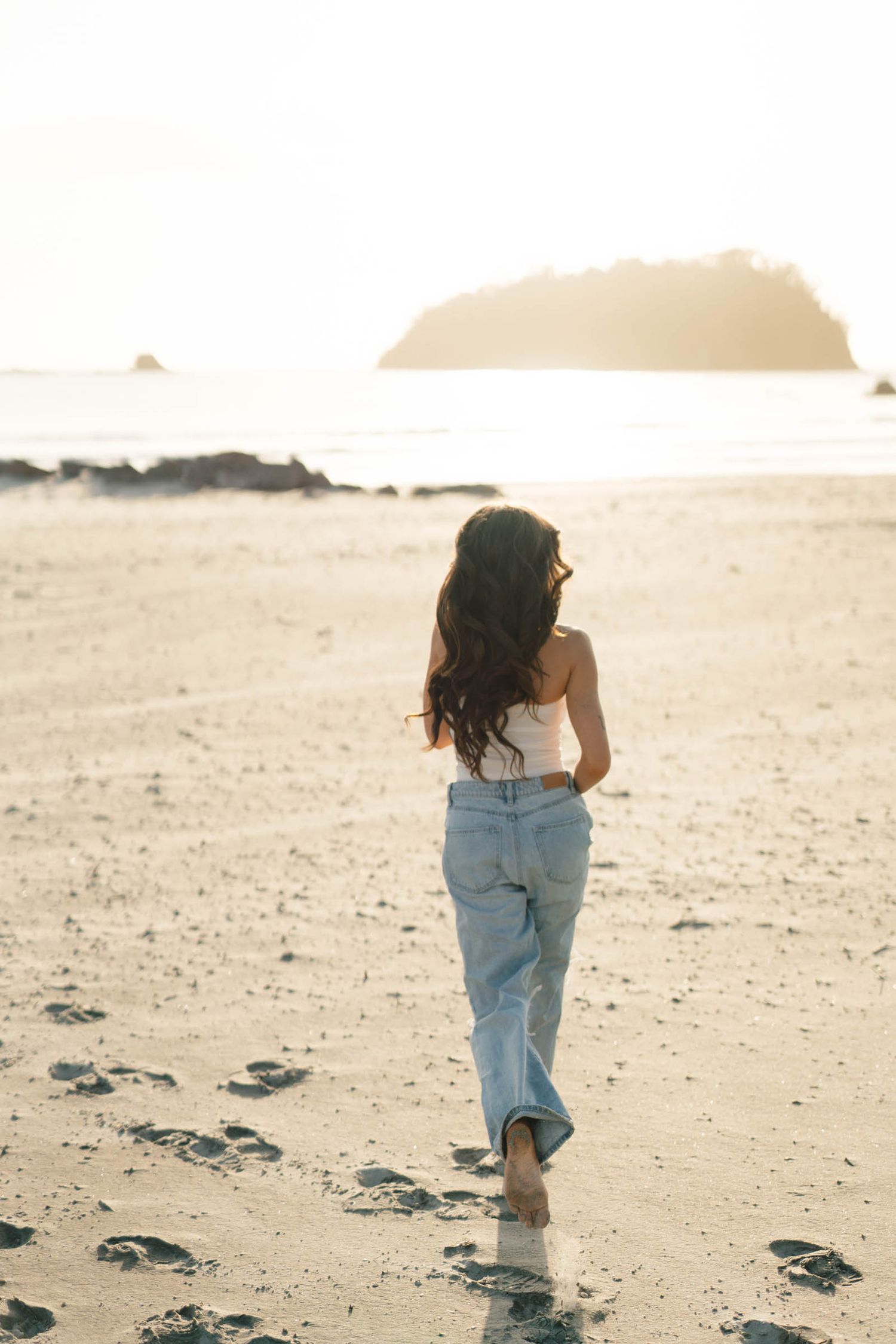 A person in jeans walks away from camera on a beach with an island silhouette in the distance.