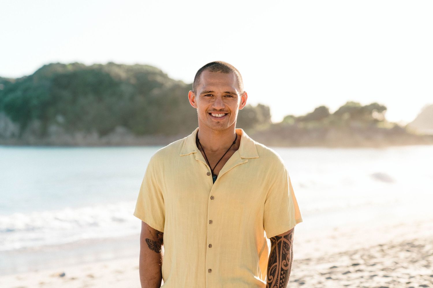 A smiling person in a light yellow shirt poses confidently during a sunny beach photoshoot.