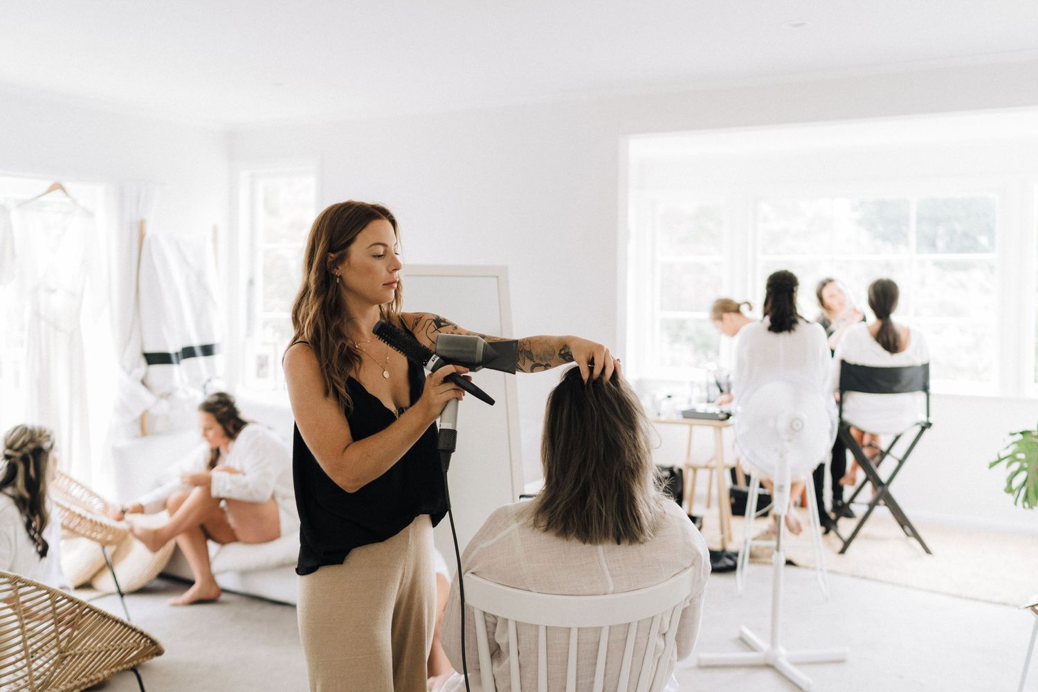 Hairstylist styling client's hair in bright white modern salon setting with other customers visible in background.