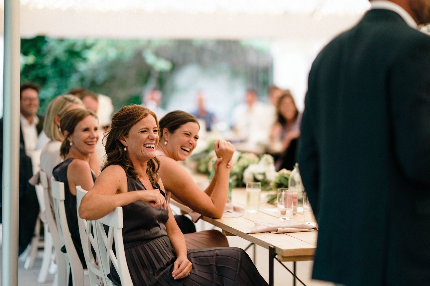 Wedding reception guests seated at long tables under a tent laughing and enjoying dinner service outdoors.