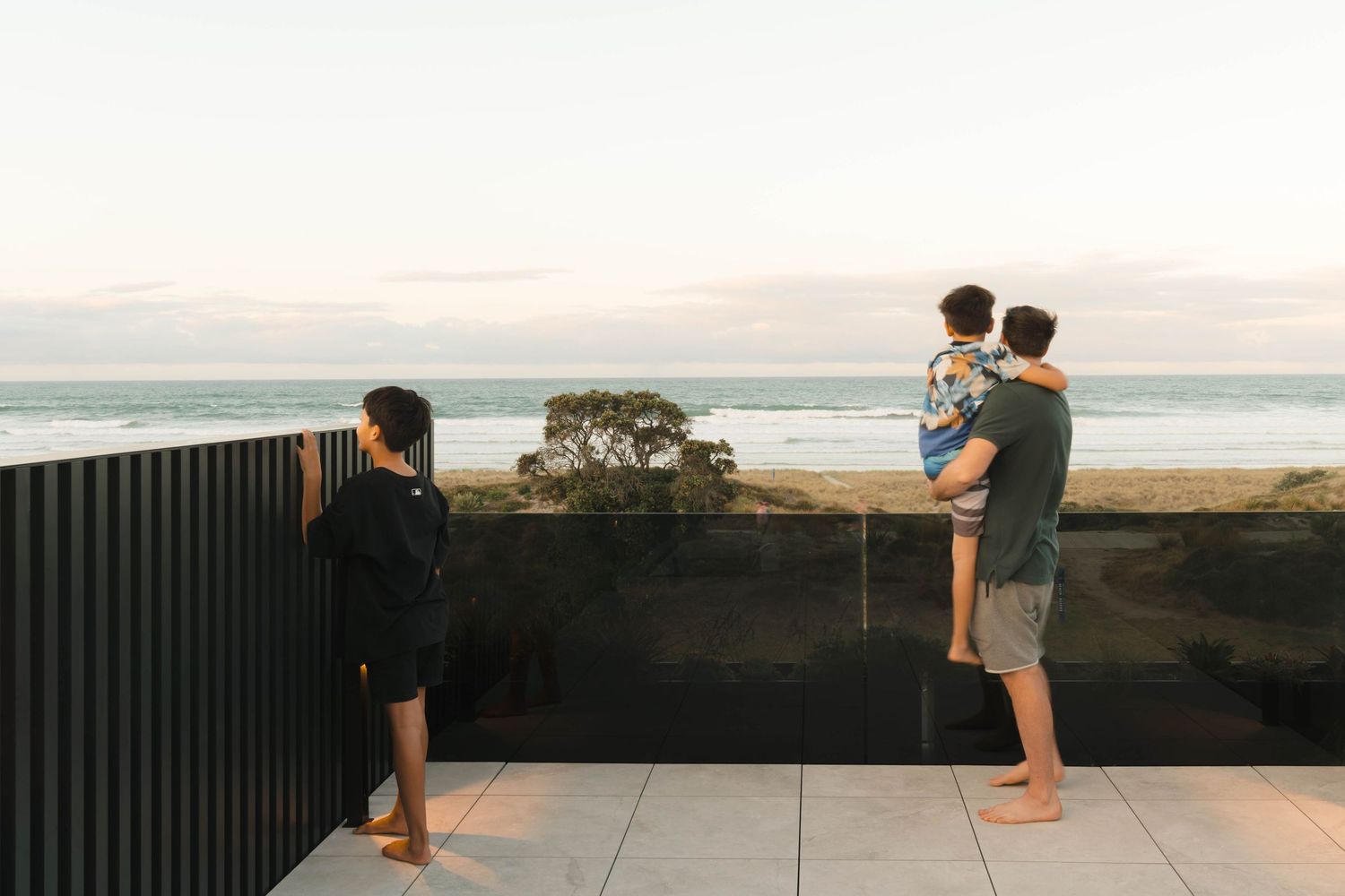 Three people standing on an oceanview balcony at sunset with a scenic coastal vista in the background.