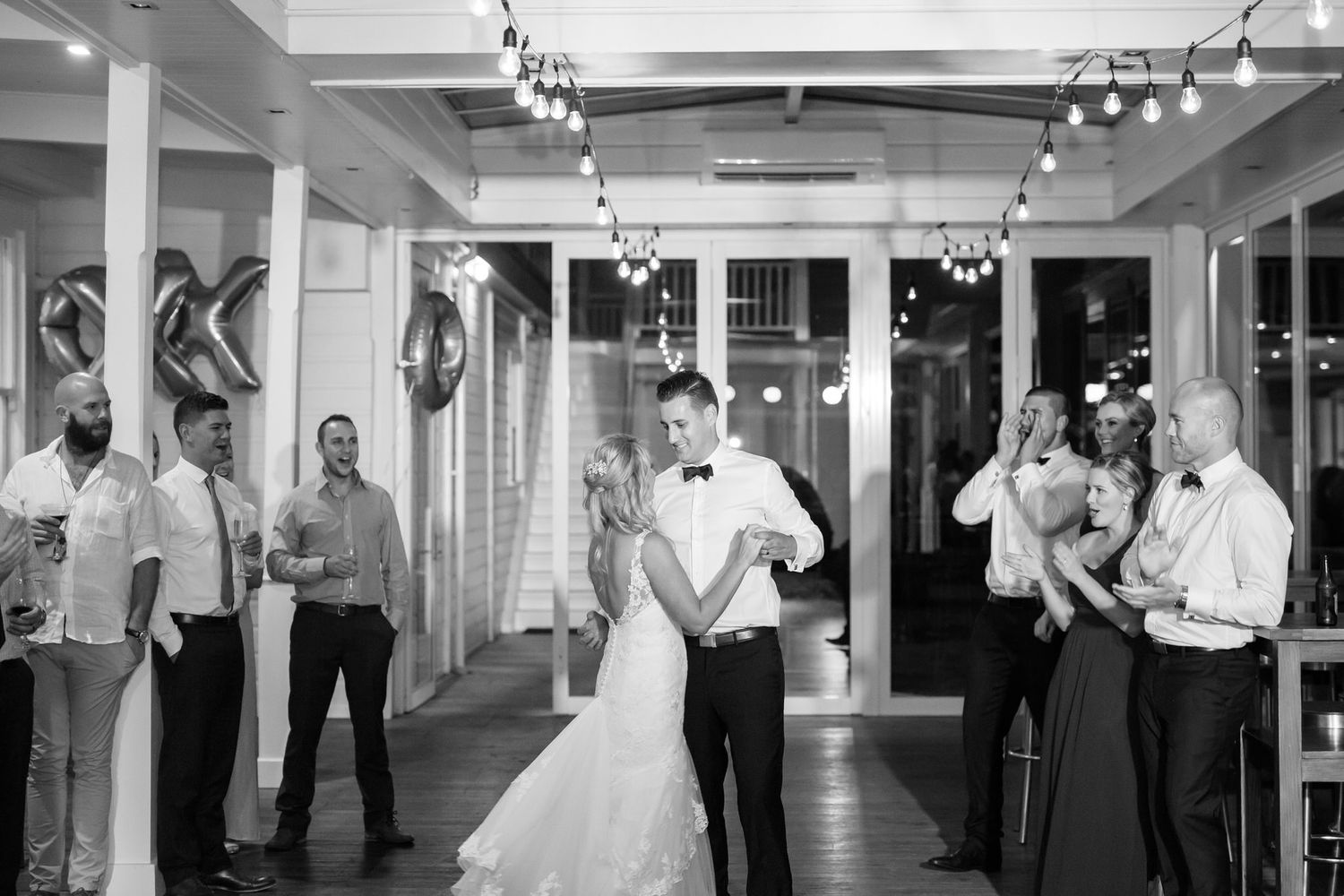Panoramic black and white photo showing wedding guests dancing in a bright indoor reception venue.
