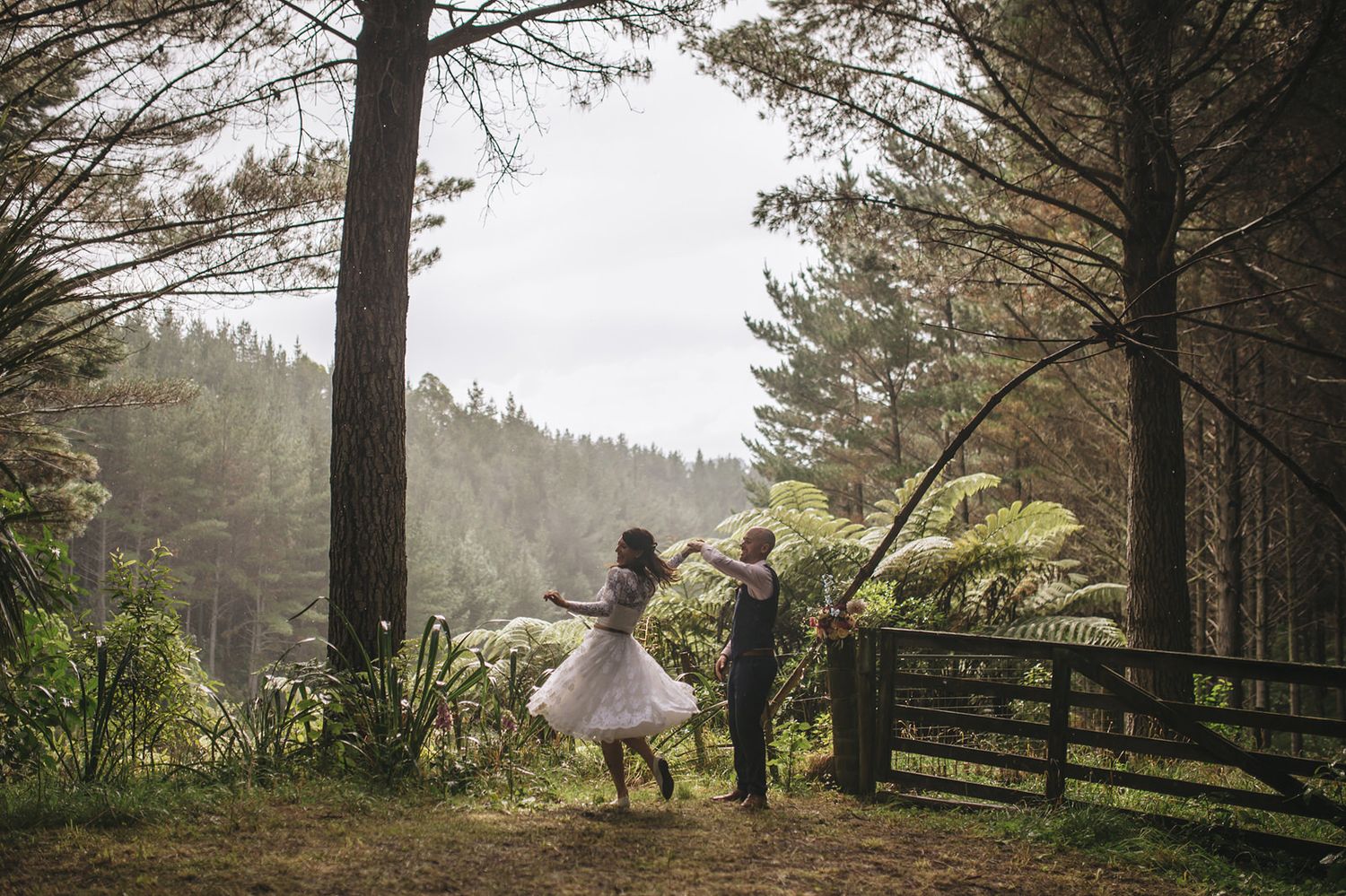 A joyful couple surrounded by tall evergreen trees dances and celebrates on a misty mountain overlook at sunset.