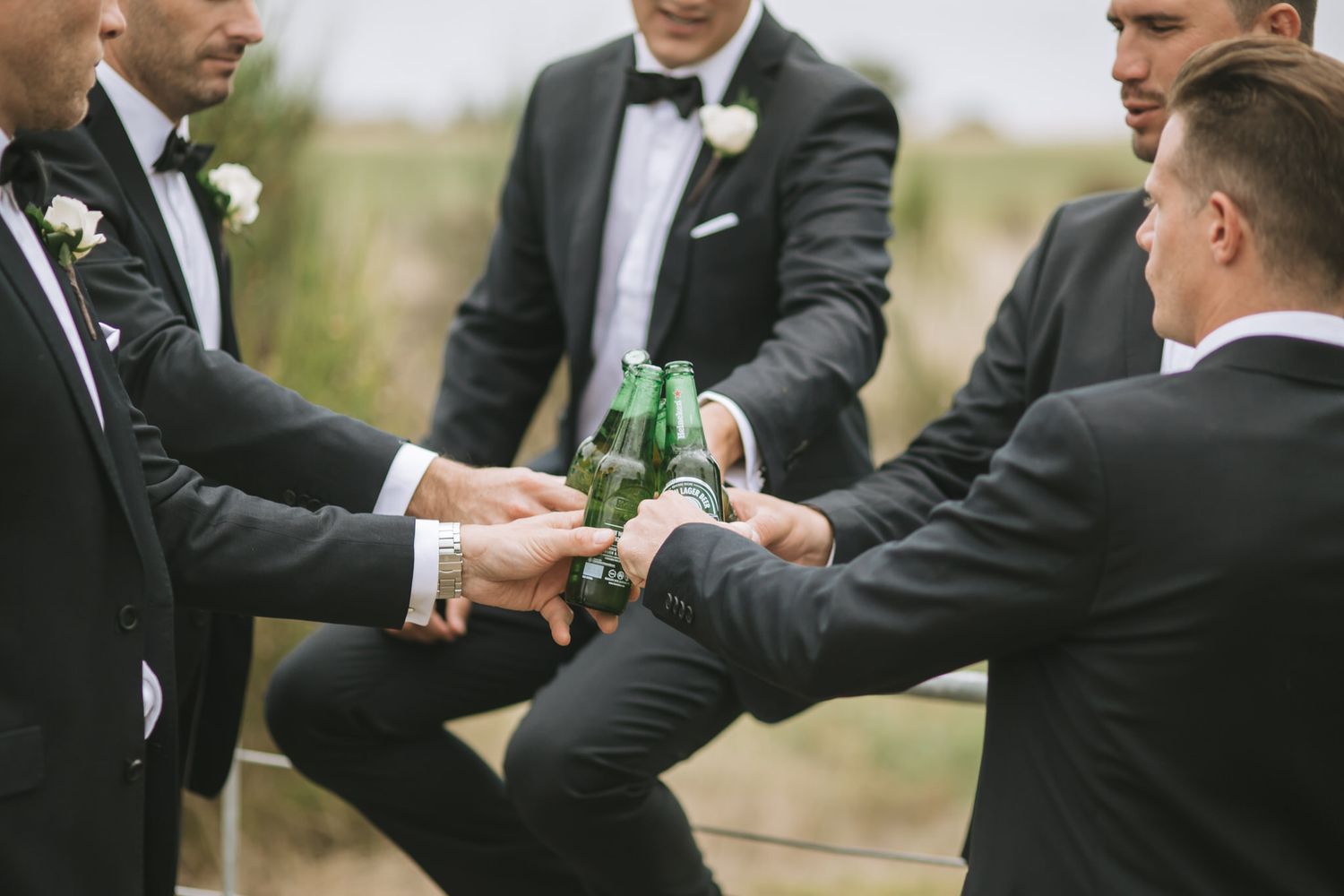 Groomsmen in black tuxedos toasting and clinking beer bottles together at outdoor wedding celebration.