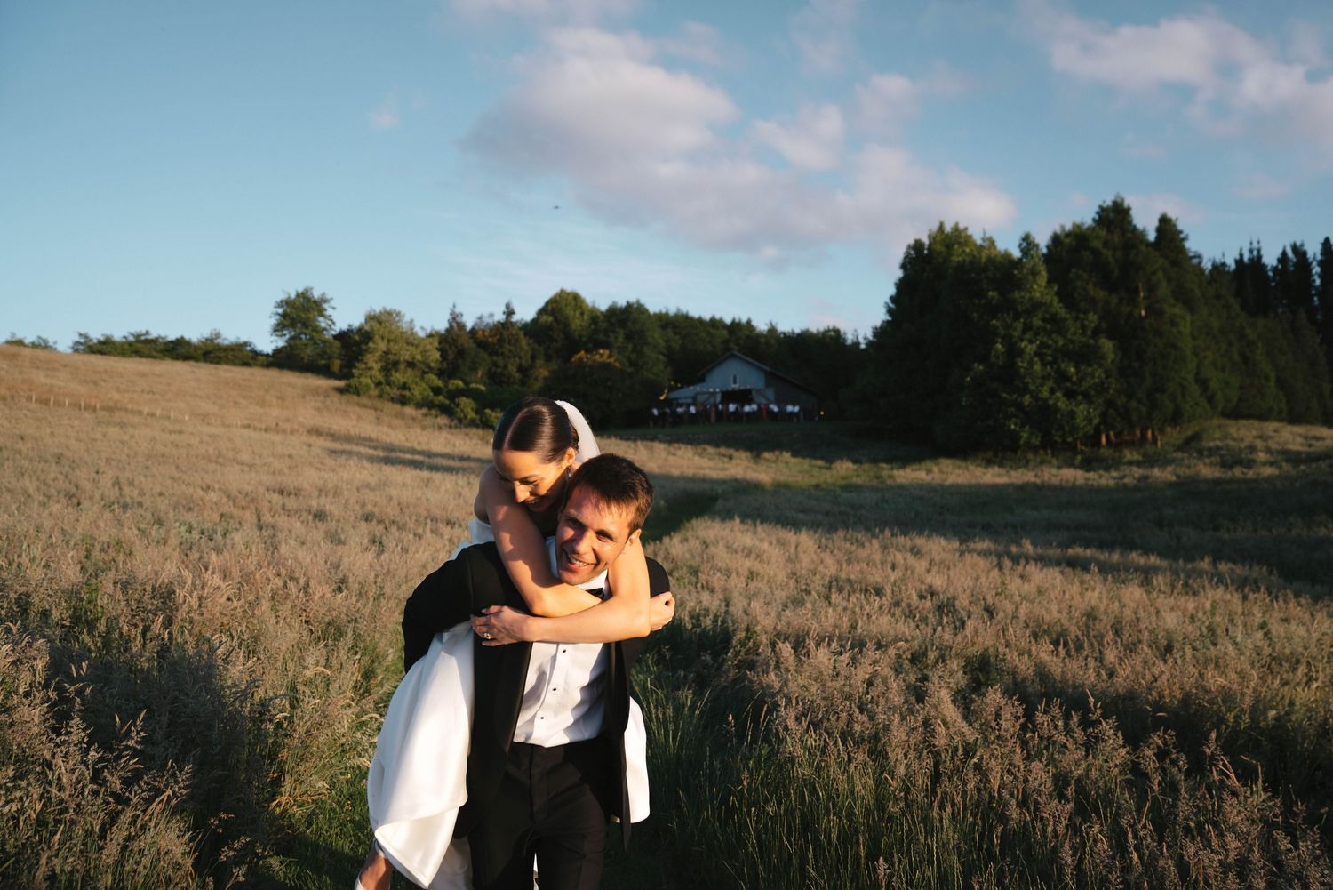 Bride and groom embracing in a sunlit meadow with trees and a white building in the background.