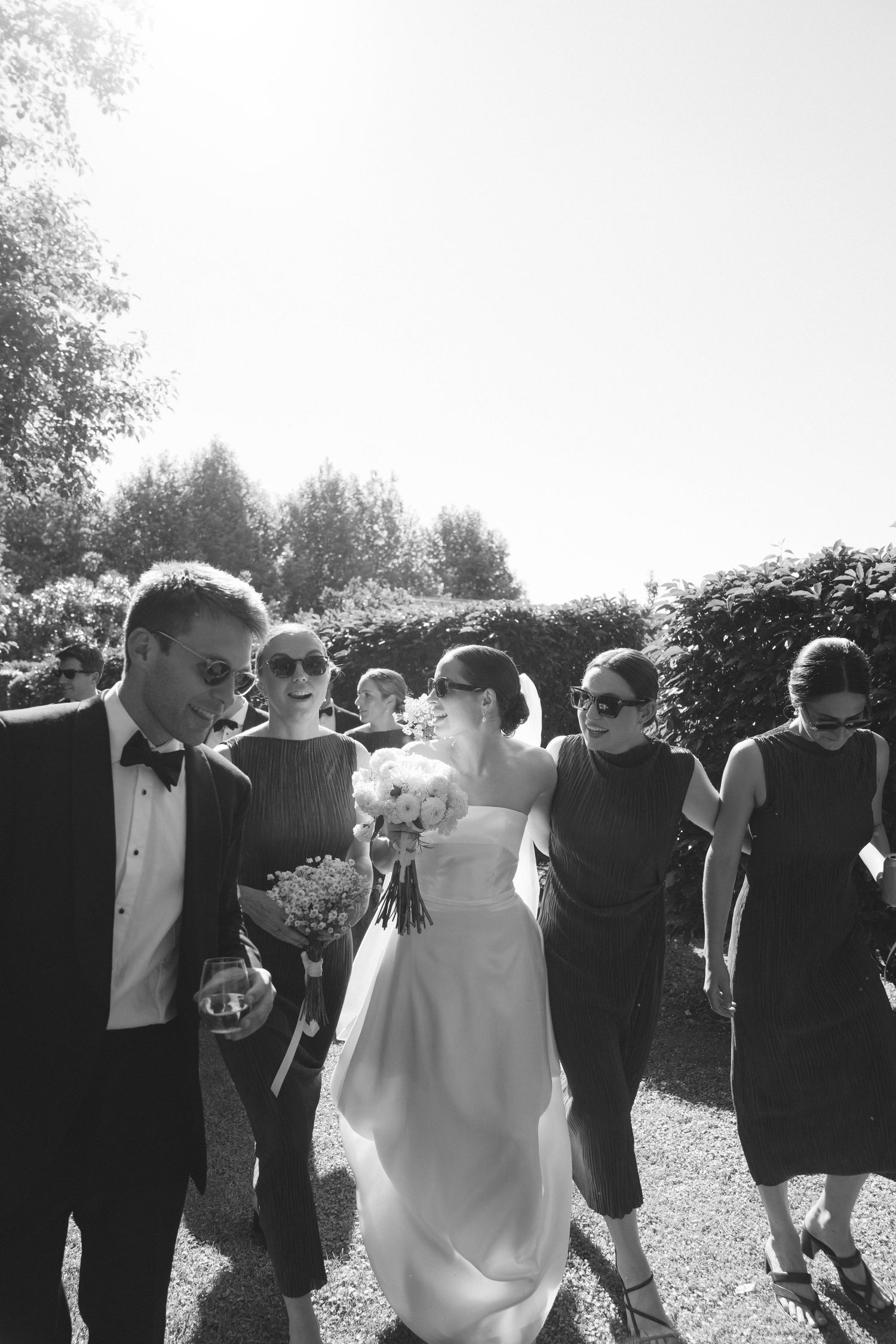 Black and white photo of a bride holding a bouquet walking with wedding guests outdoors on a sunny day.