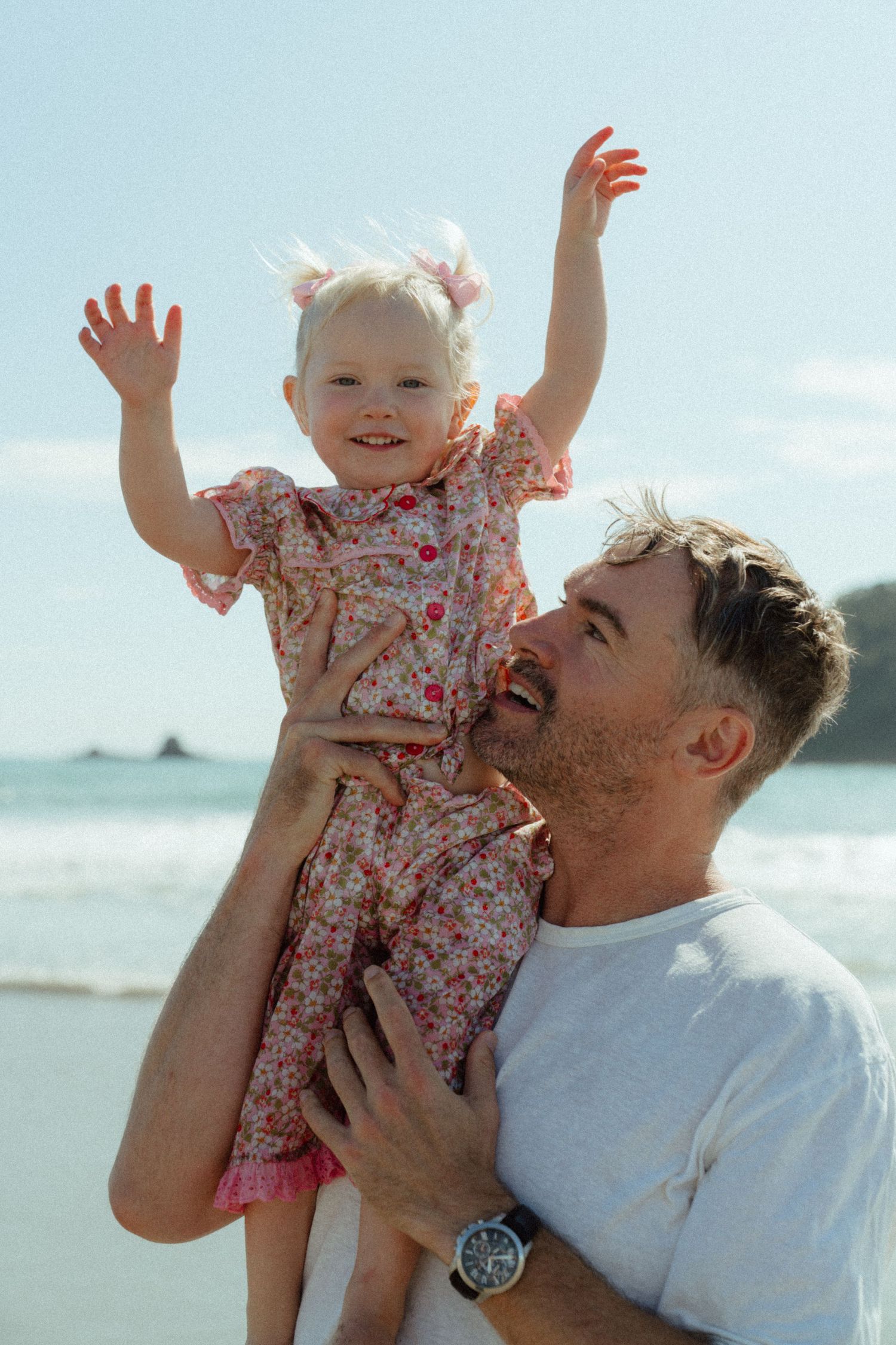 A parent lifts and plays with a small child wearing a floral dress at the beach on a sunny day.