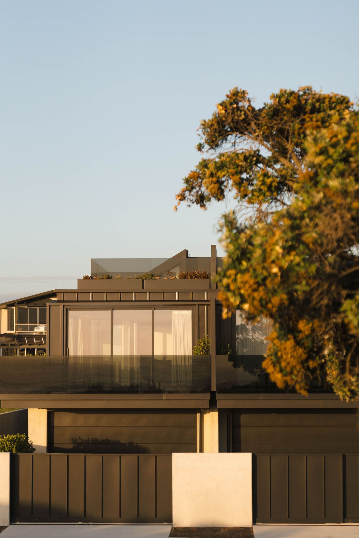 Modern two-story home with large glass windows bathed in golden sunset light, framed by lush green trees against a clear sky. Newfound.