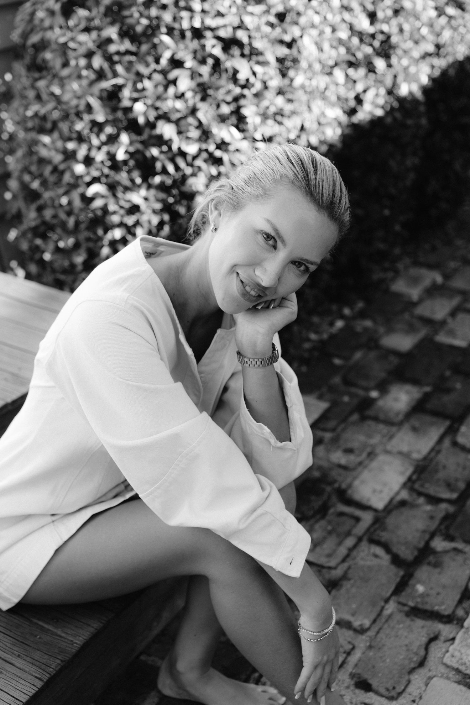 Black and white photo of a smiling woman in a white shirt sitting outdoors on a wooden deck near a brick wall.