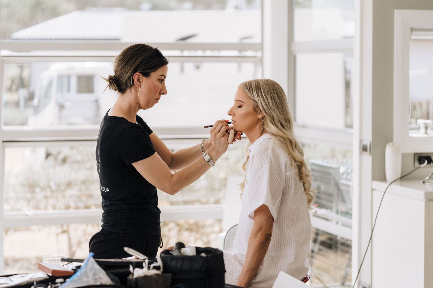 A makeup artist applies cosmetics to a client in a bright, window-lit setting during a professional beauty session.