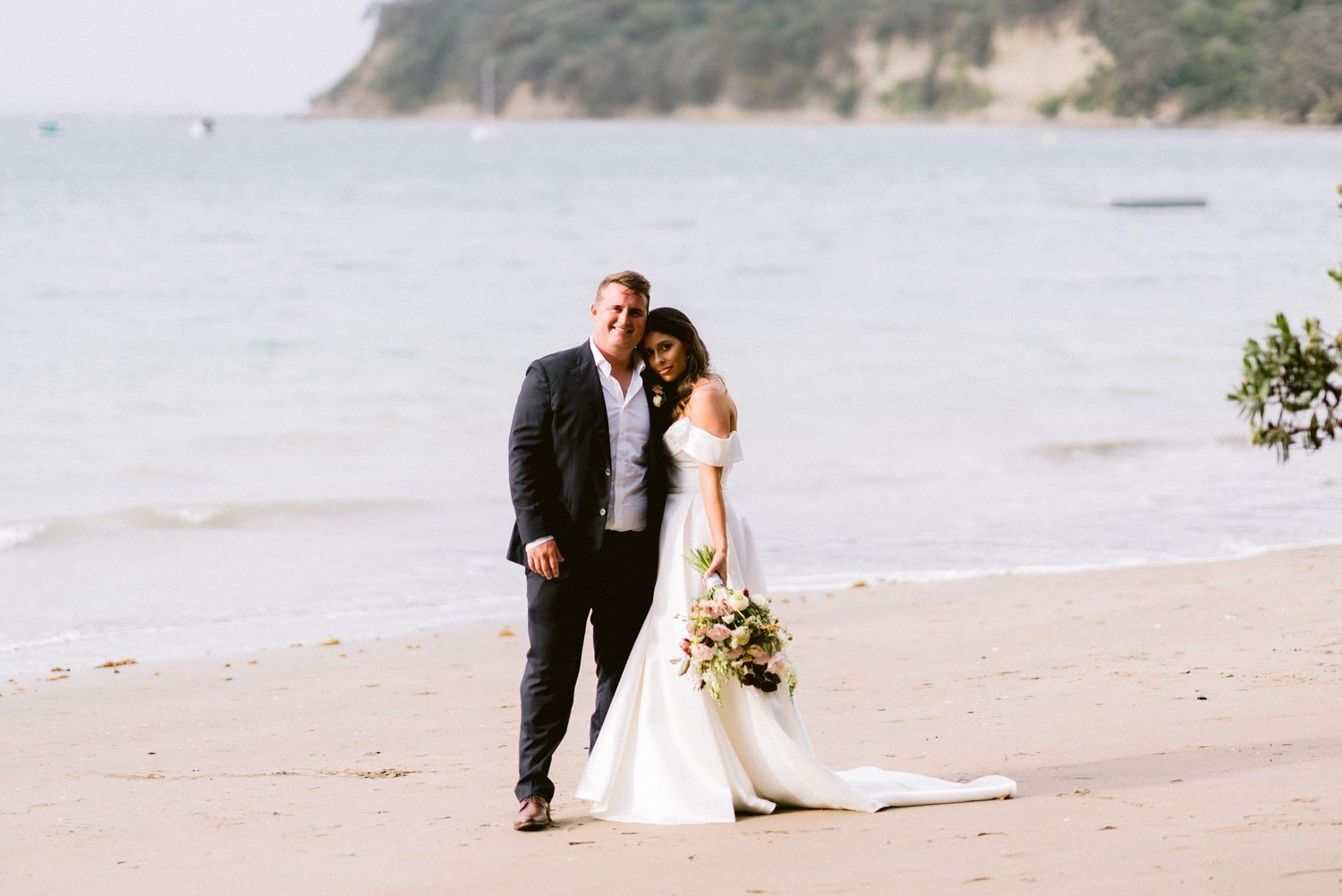 A couple poses for wedding photos on a scenic beach with cliffs in the background.