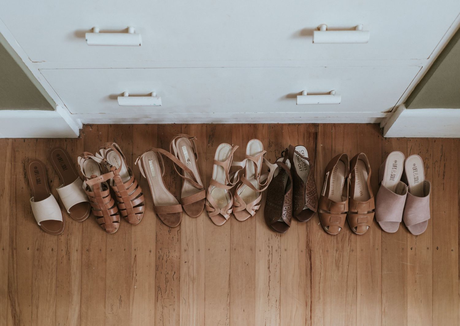 A row of assorted shoes and boots lined up neatly against a wooden floor and white wall.