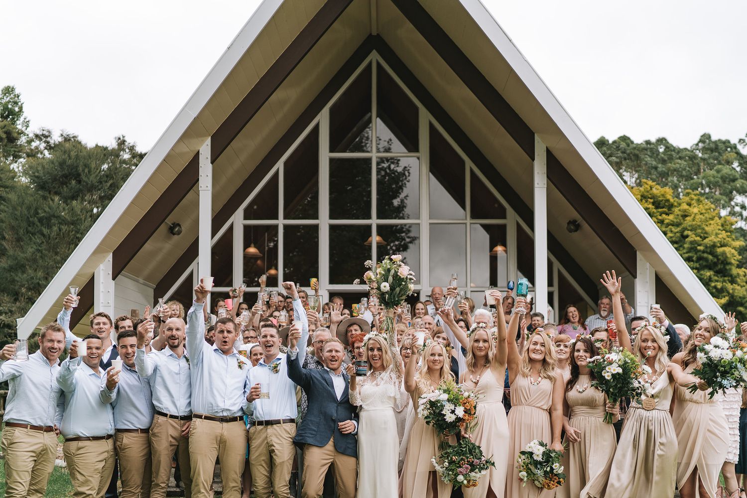 A group in coordinated pastel and khaki wedding attire poses for photos in front of a white A-frame church.