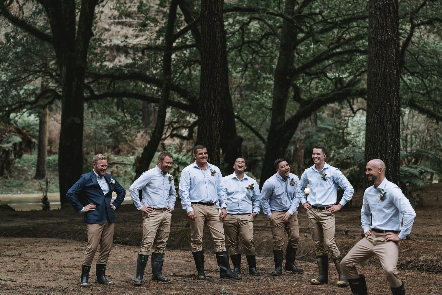 A group of people in light blue shirts and khaki shorts gather casually on a woodland path among tall trees.