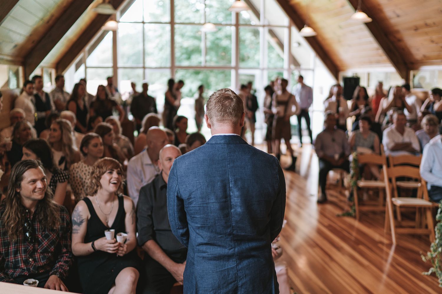 Wedding attendees gather inside a rustic wooden venue hall with exposed beams and natural lighting.