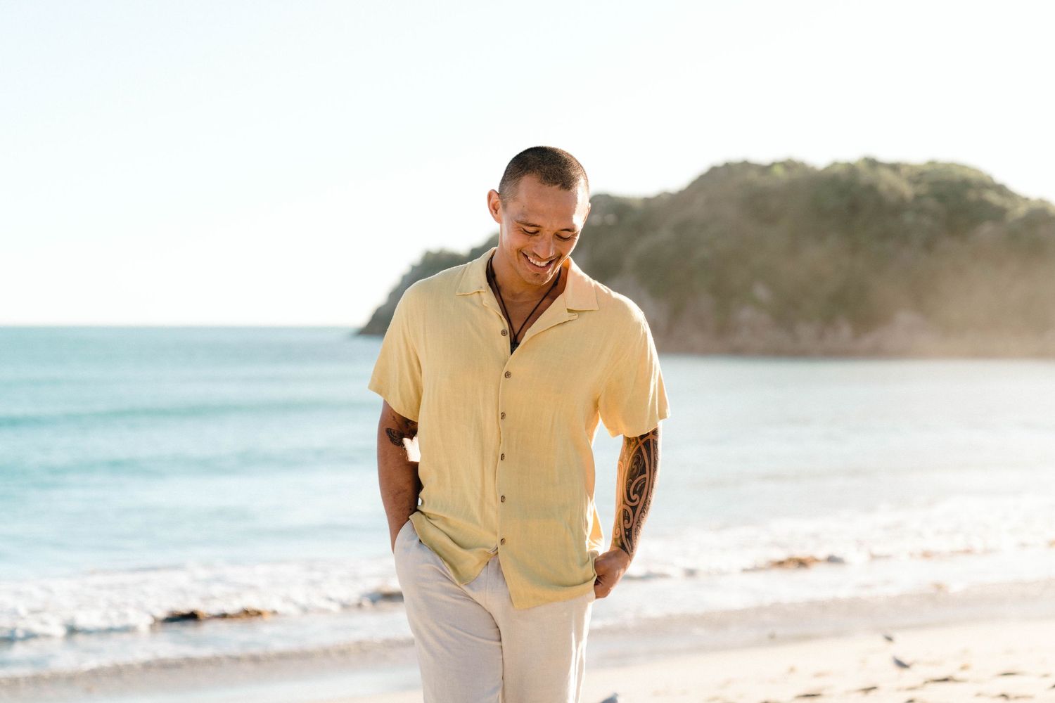 Someone wearing a yellow button-down shirt smiles while walking on a sunny beach with cliffs in the background.