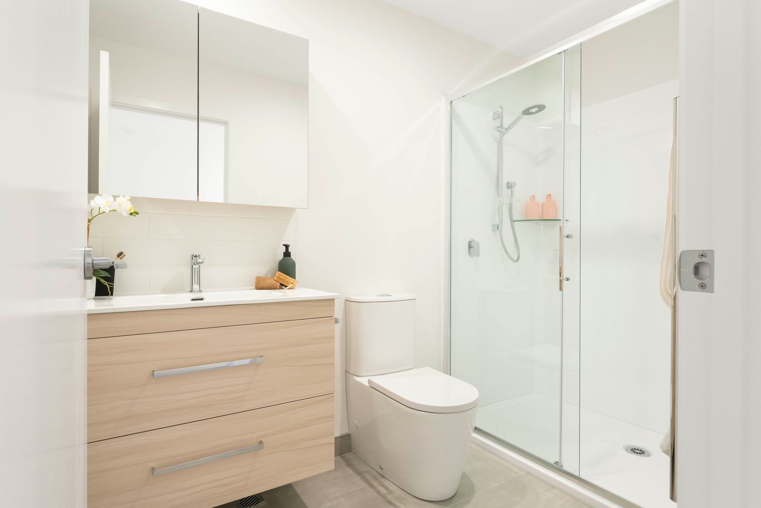 Modern minimalist bathroom interior with white walls, large mirror, wooden vanity and glass shower enclosure.