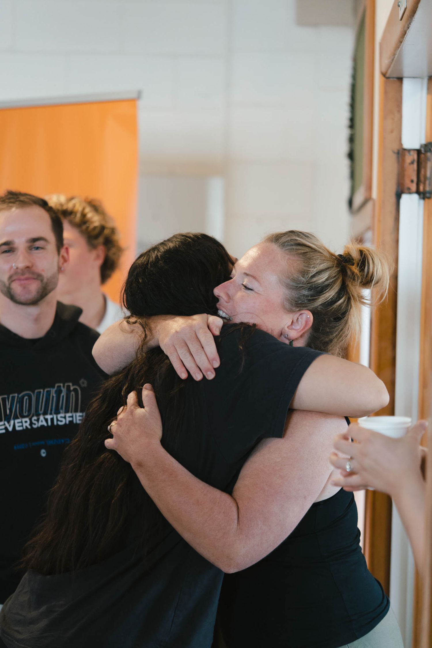 People embracing and hugging in an indoor setting with orange walls visible in the background.