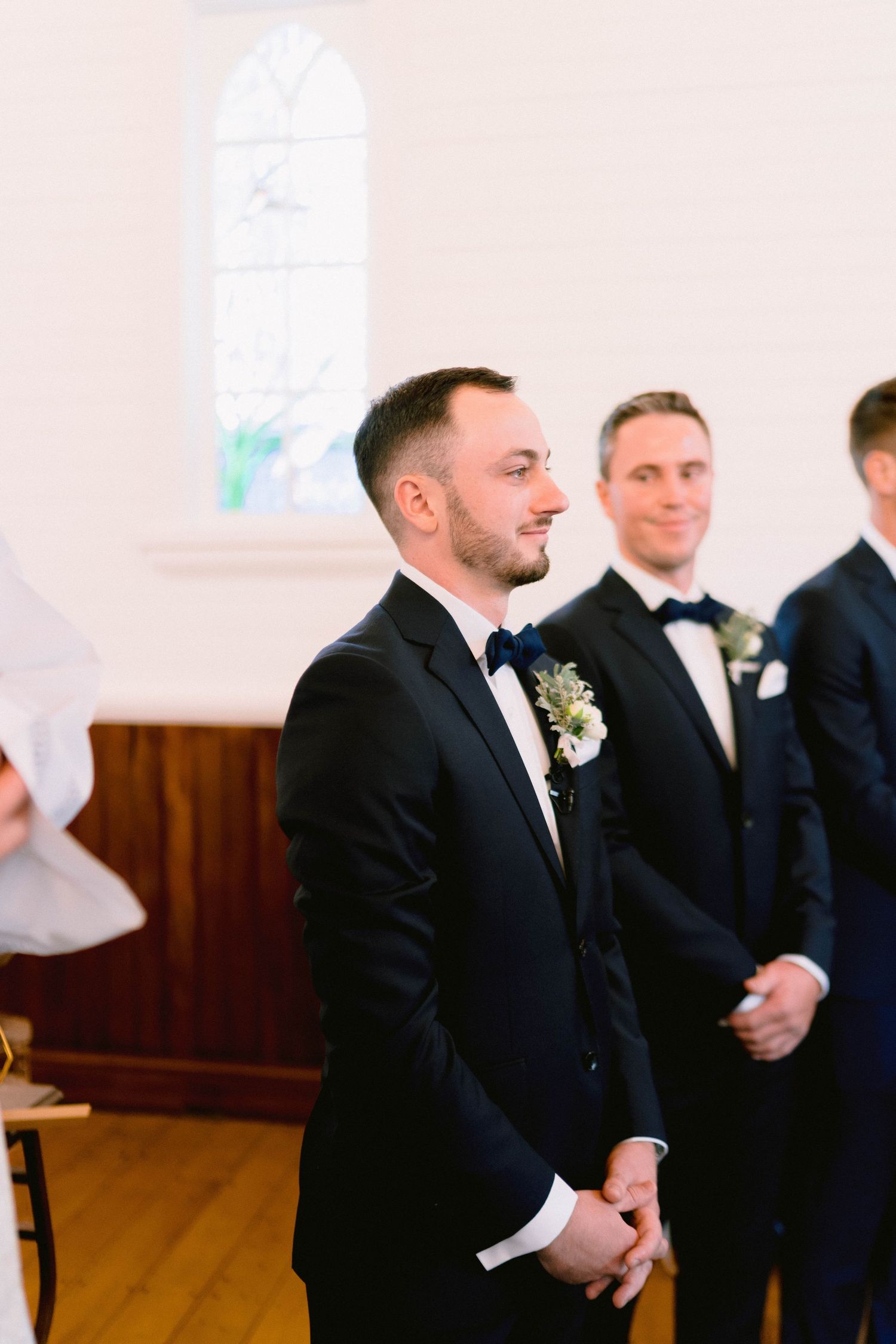 A groomsman smiles during an elegant church wedding ceremony with a white arched window in the background.