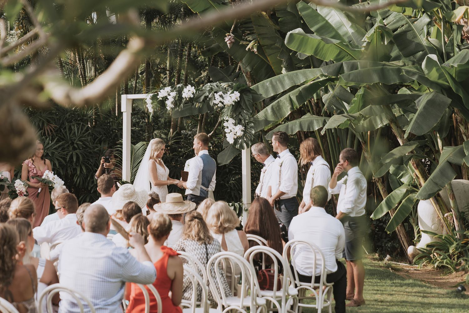Guests gathered at an outdoor wedding ceremony with tropical garden setting and white decor.