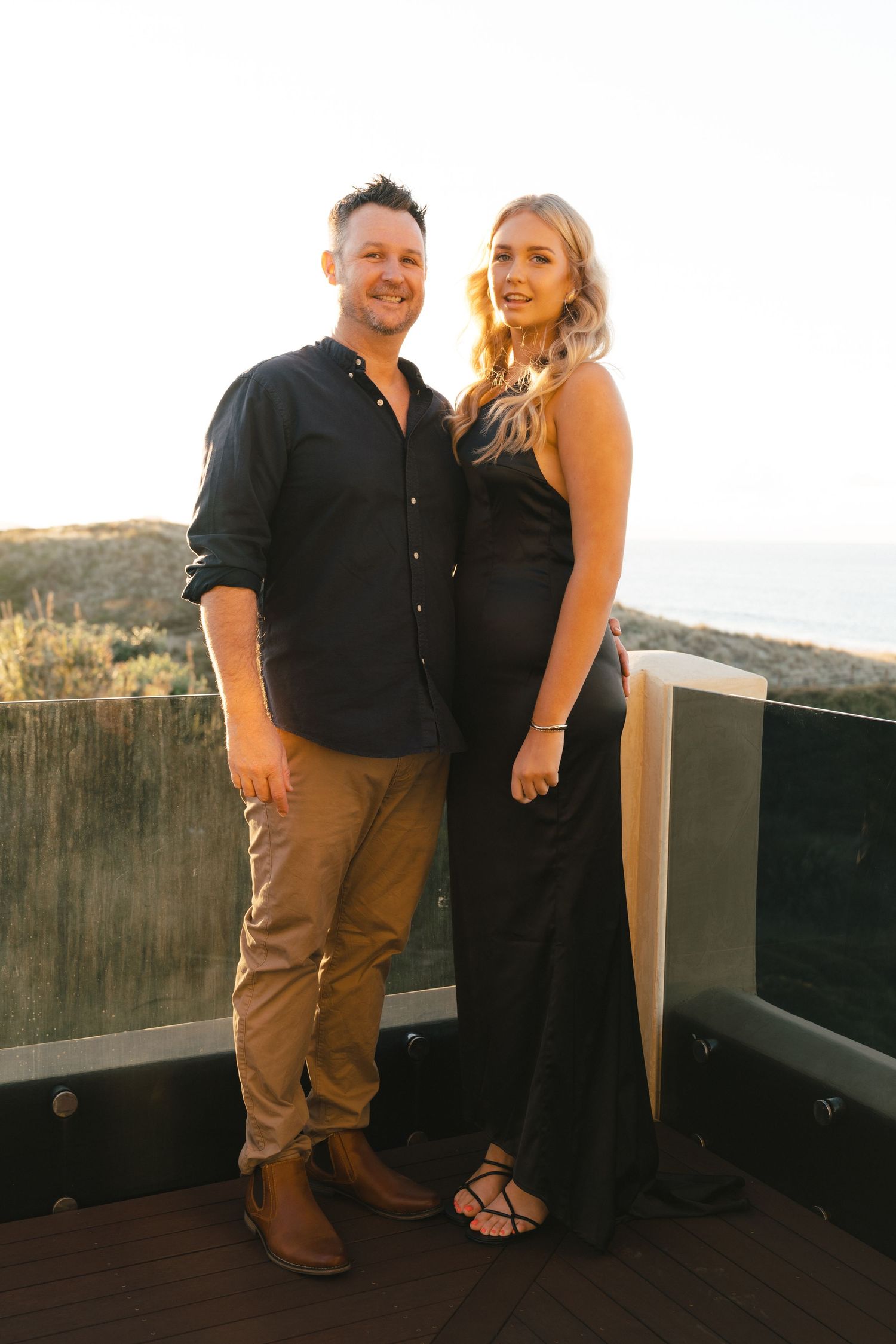Couple posing together on wooden deck during golden hour in coordinated black outfits.
