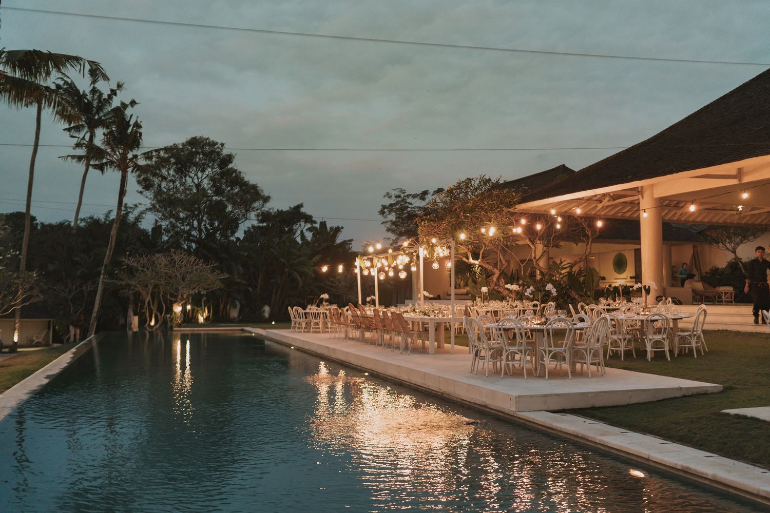 Evening poolside wedding reception setup with string lights and white dining tables.