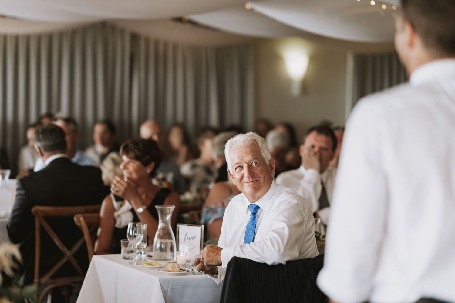 Wedding guests seated at tables during reception while a server in white tends to diners in an elegant venue.