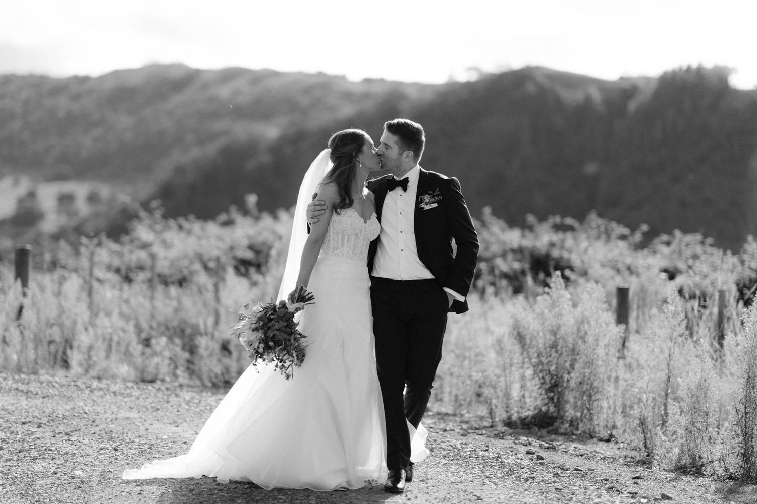 Black and white outdoor wedding photo showing couple embracing on rural mountain path with scenic backdrop.