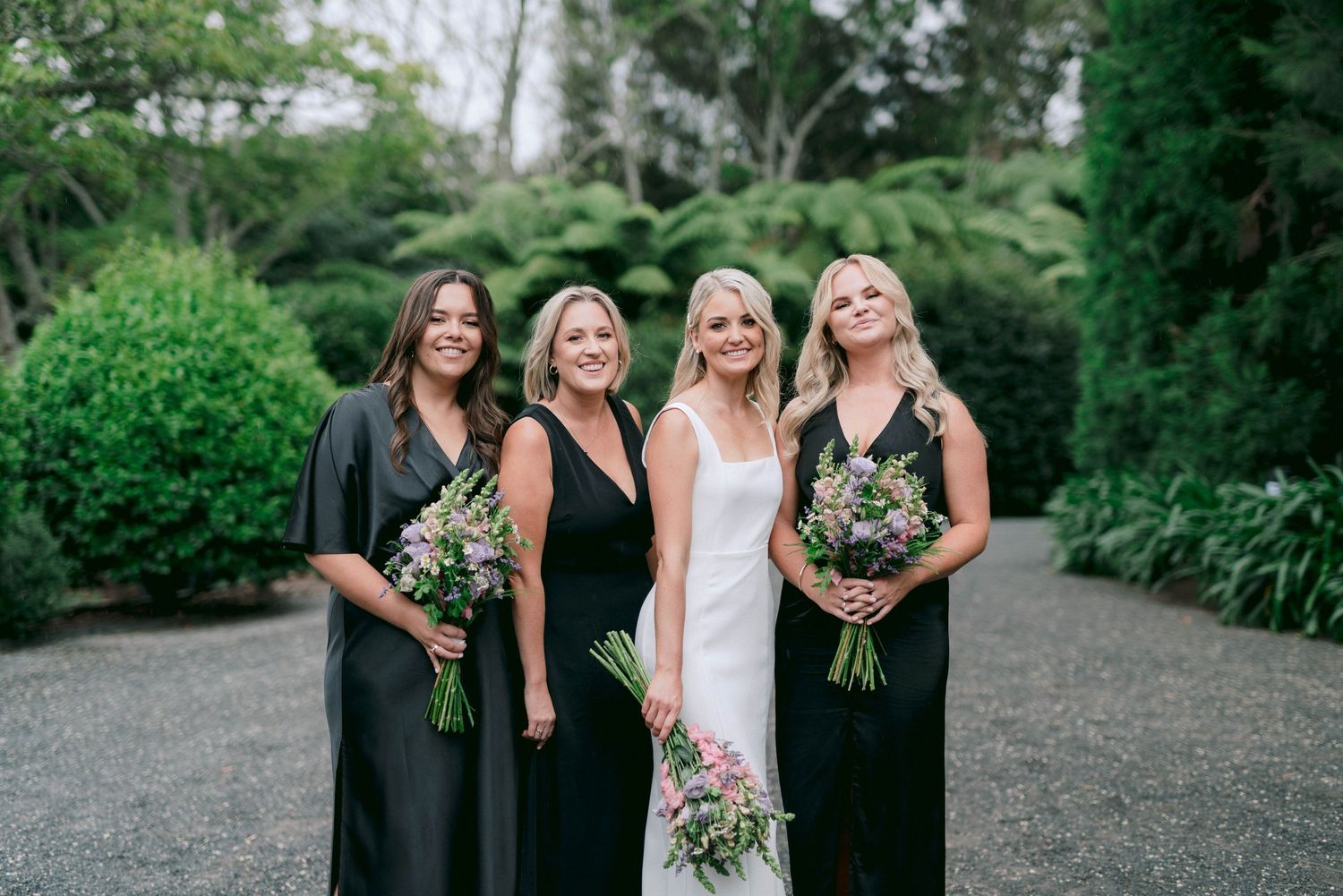 Four women in formal attire stand together holding floral bouquets on a garden path.