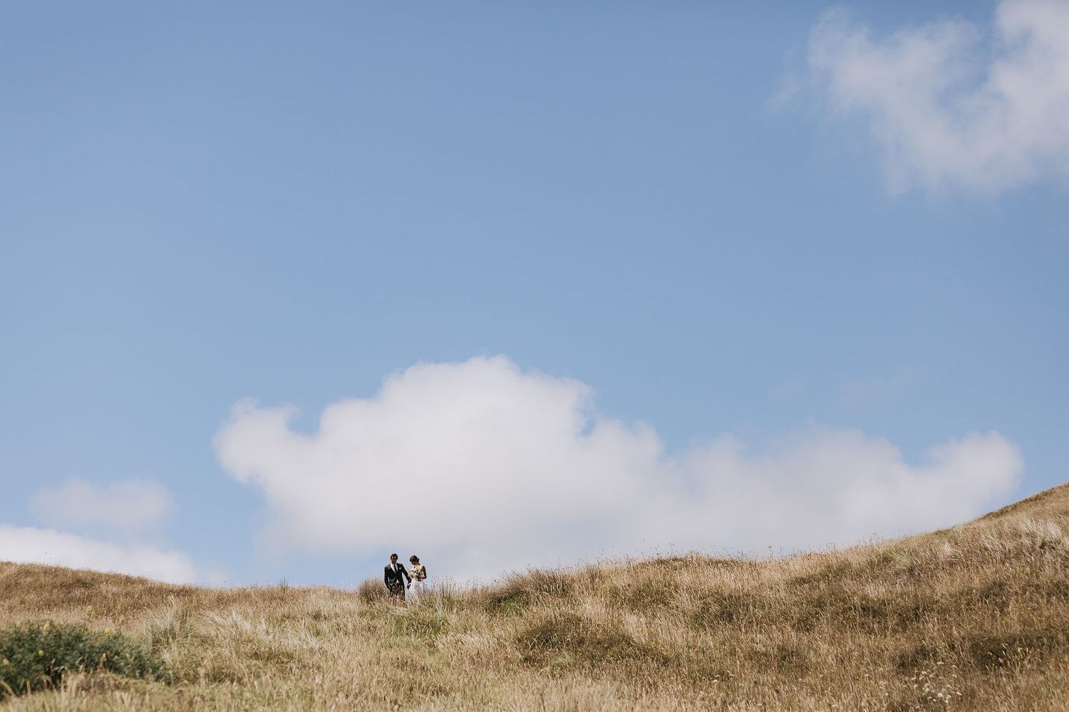 Two hikers stand atop a grassy hill against a blue sky with white puffy clouds.