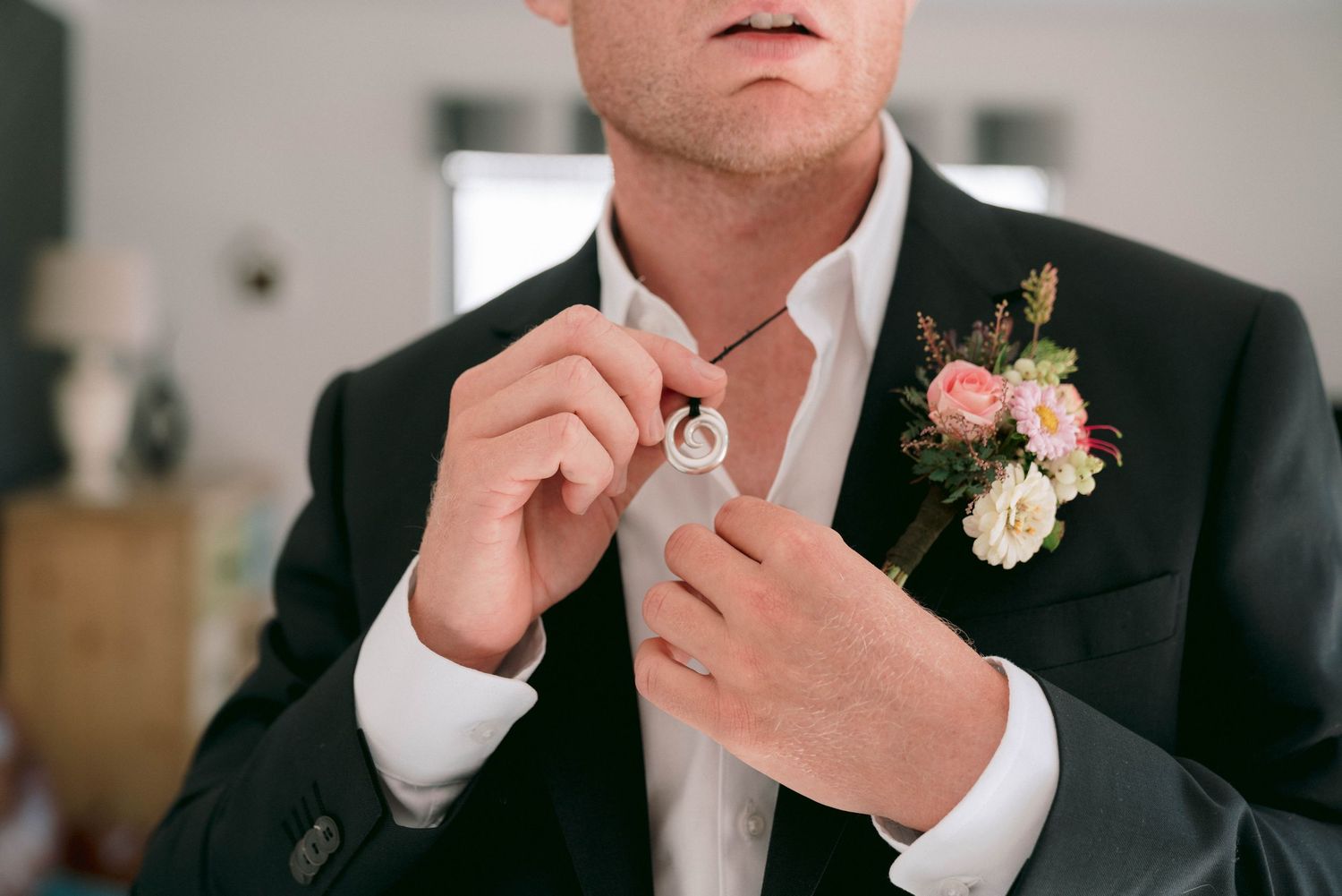 A person in a black suit adjusts their tie while wearing a pink floral boutonniere.