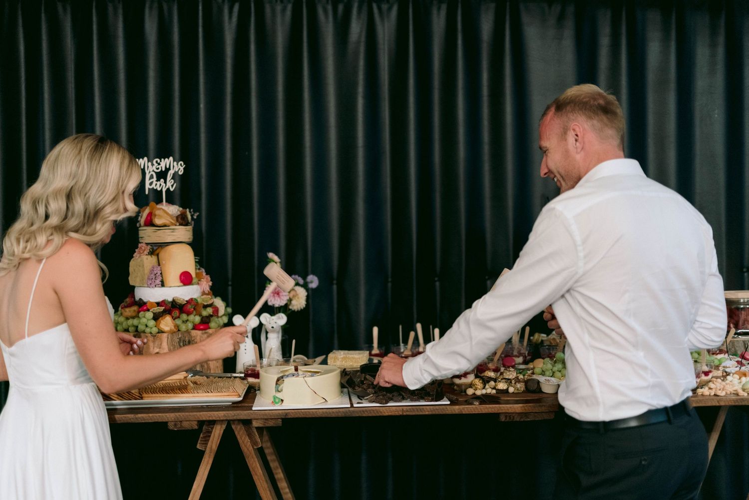 A couple cutting their wedding cake together at a rustic style dessert table with dark curtain backdrop.