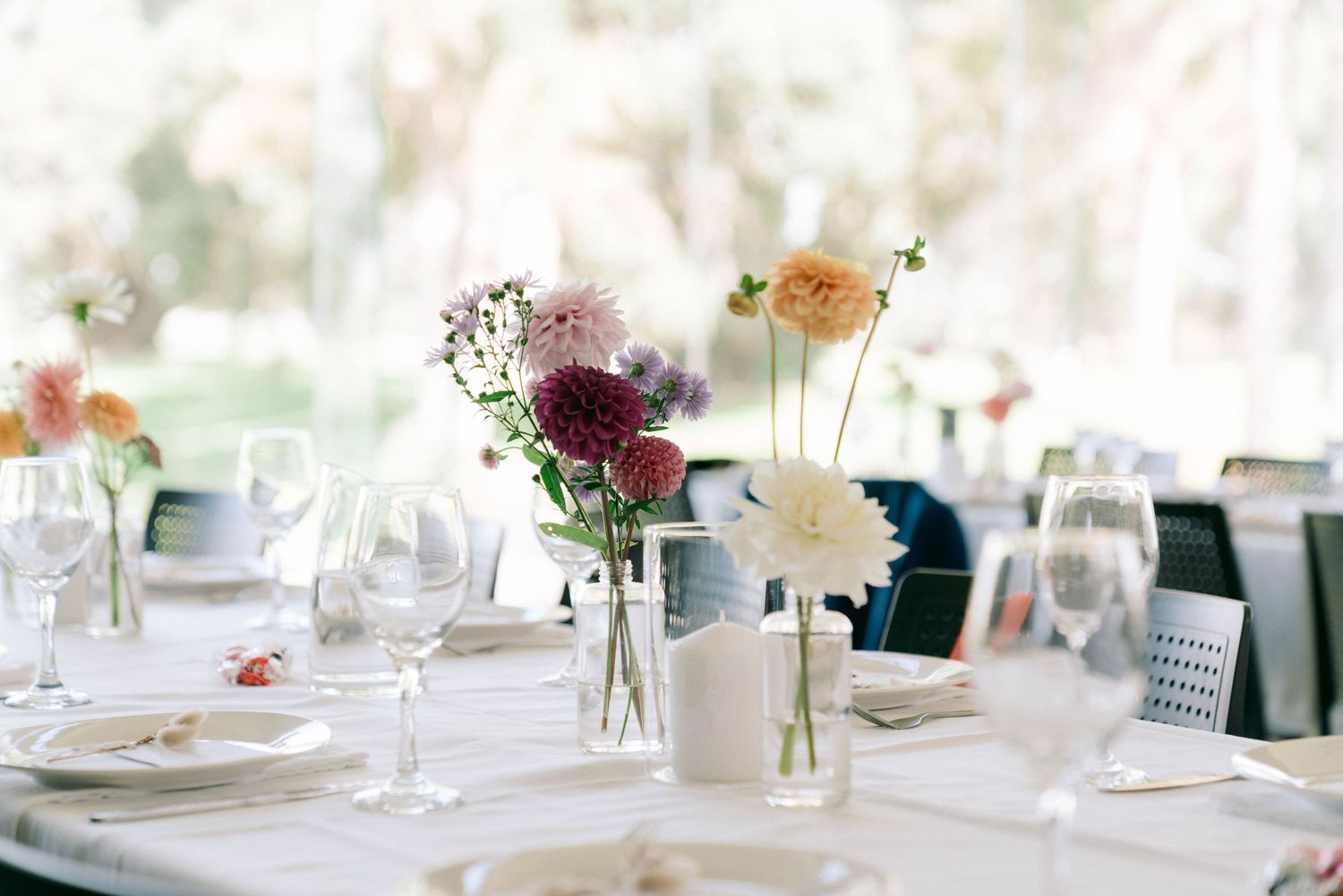 An elegant table setting at a wedding reception with white linens, wine glasses and delicate floral centerpieces in glass vases.