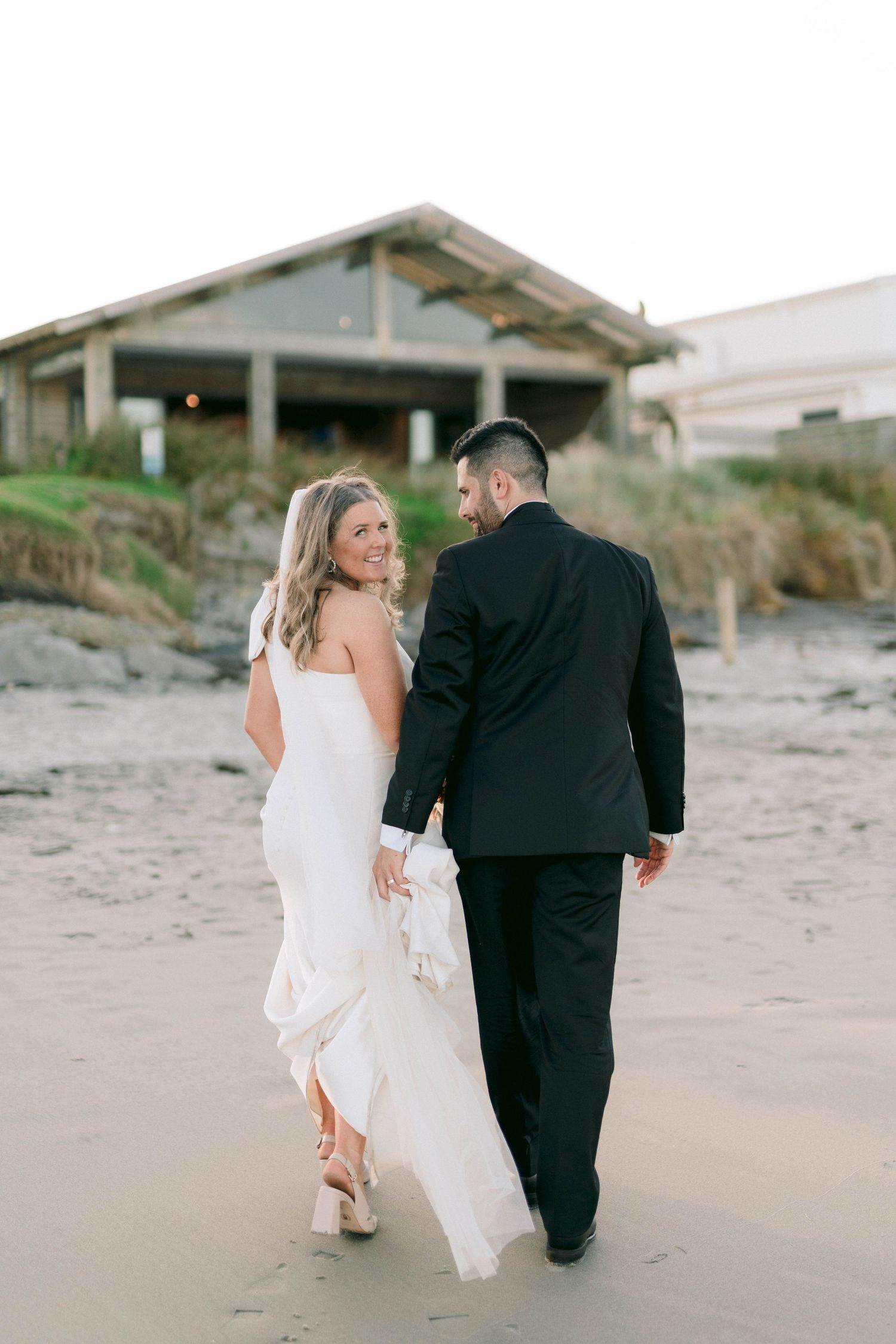 Couple walks hand in hand along sandy beach with rustic beach houses in background at dusk.
