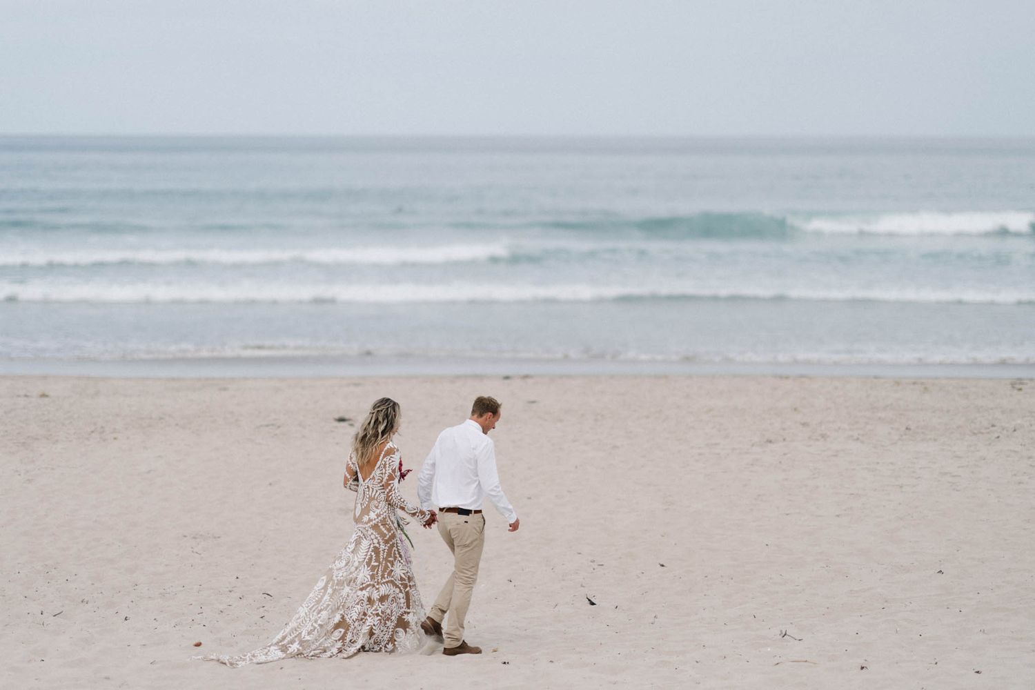 Two people walking along a misty oceanfront beach with rolling waves and sand extending into the distance.