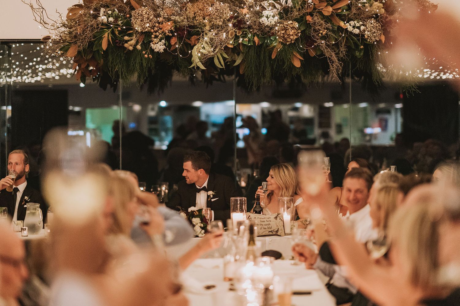 Wedding guests seated at reception dinner tables beneath elegant floral arrangements and warm ambient lighting.