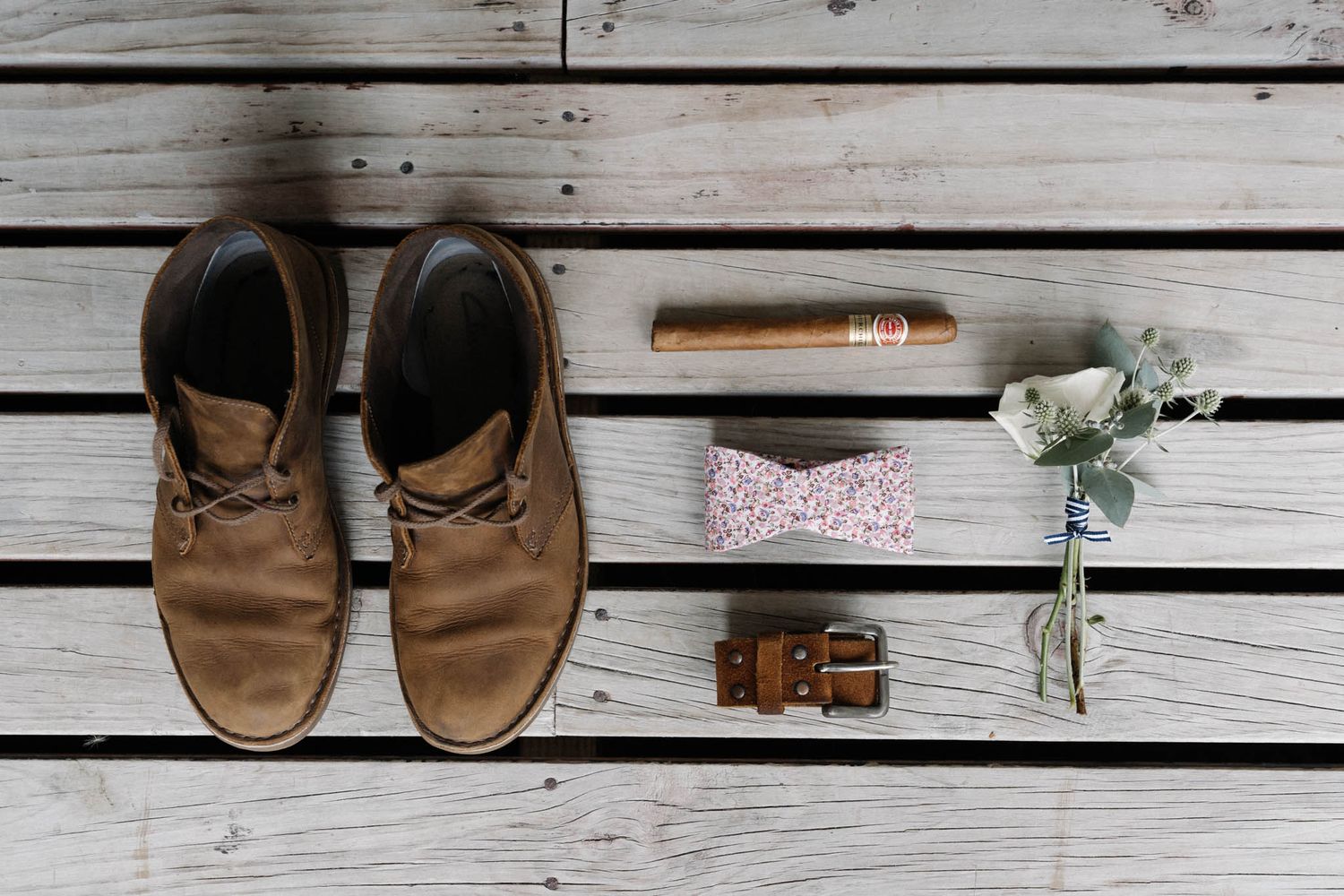 Flat lay of brown leather boots, belt, flowers and pink hair accessory arranged on white wooden planks.