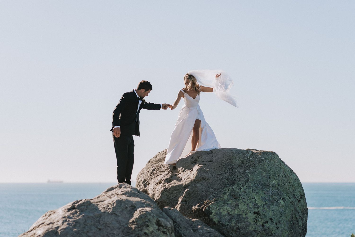Romantic newlyweds stand together atop a dramatic seaside cliff during their ocean view wedding photoshoot.