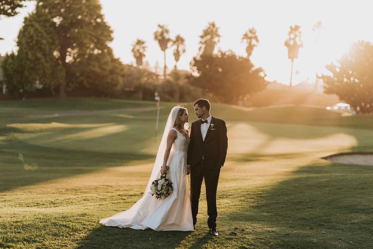 A couple stands together on a sunlit golf course during golden hour with palm trees and rolling greens in the background.