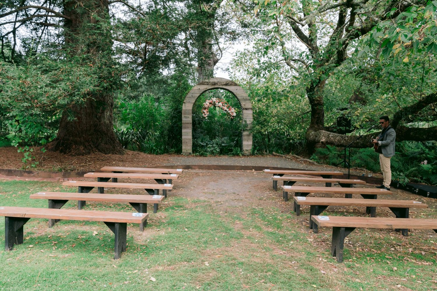 Wooden benches arranged in rows beneath towering trees create an intimate outdoor ceremony space.