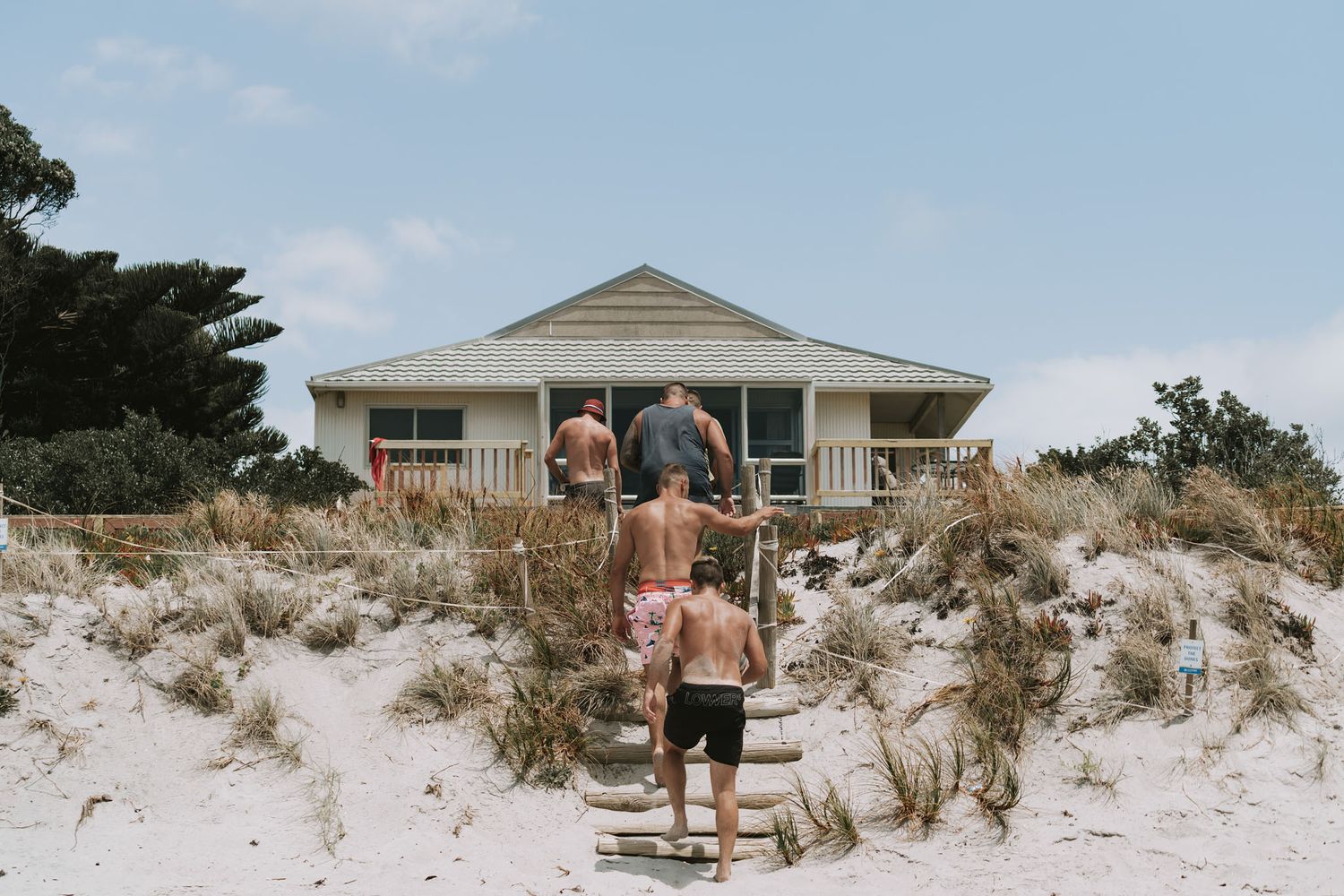 People at a beach house with a sandy pathway through coastal dunes leading to a beige cottage on a sunny day.