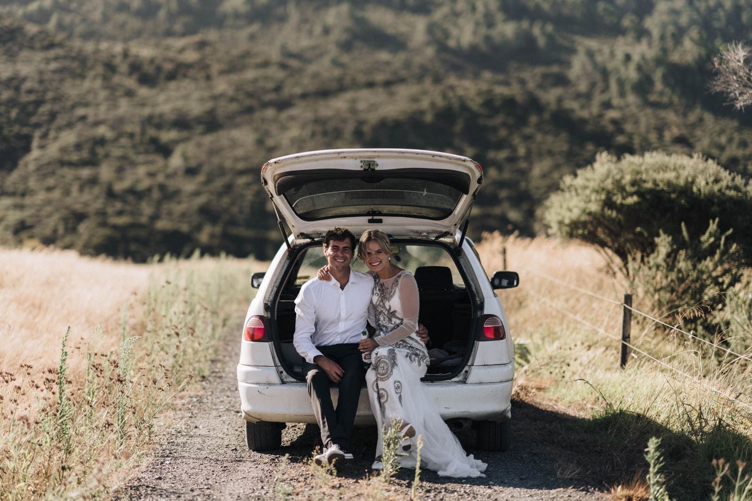 A couple sits together on the open trunk of a car parked on a scenic mountain road in summer.