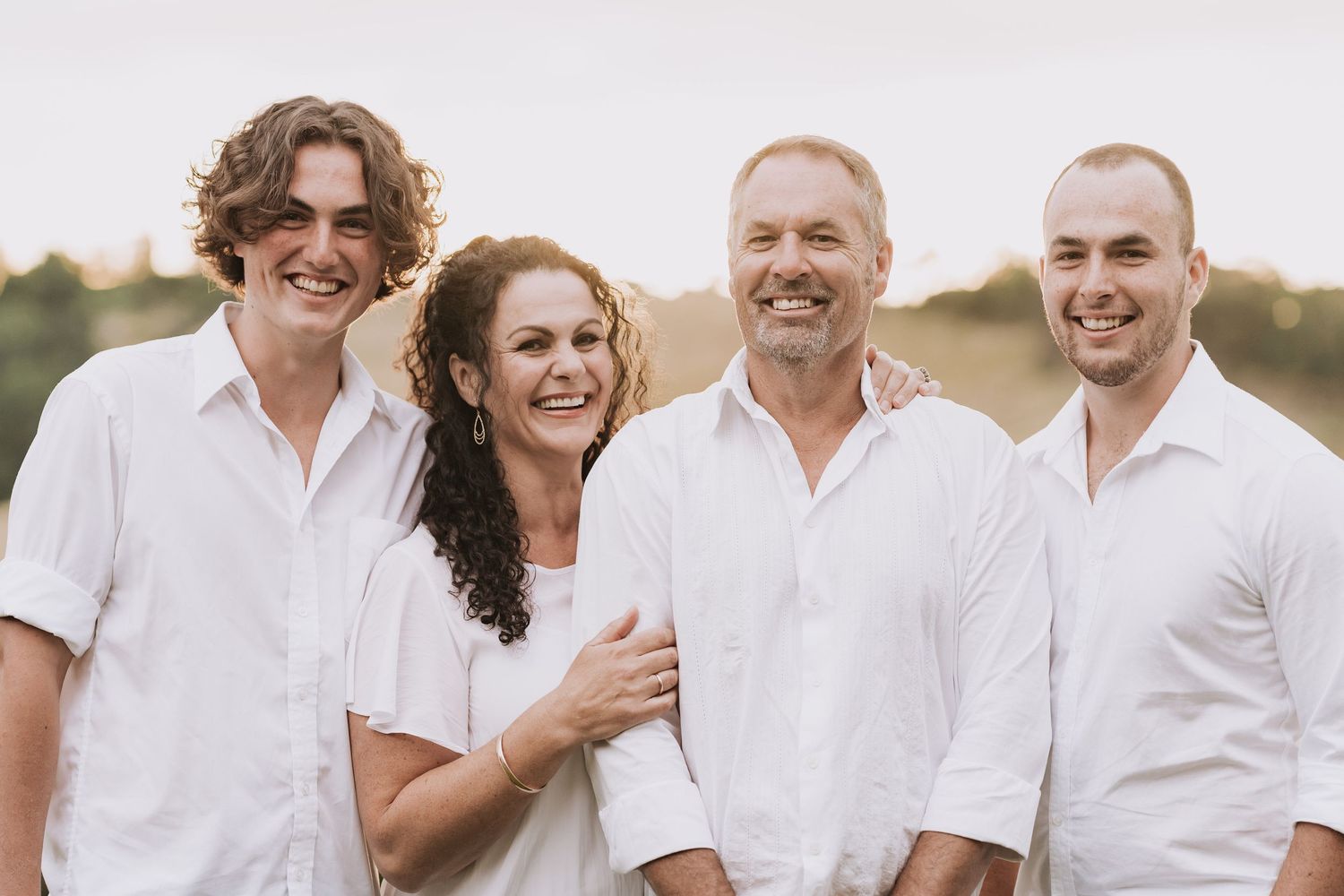 Family wearing coordinated white outfits laughing and embracing together outdoors during golden hour sunset.