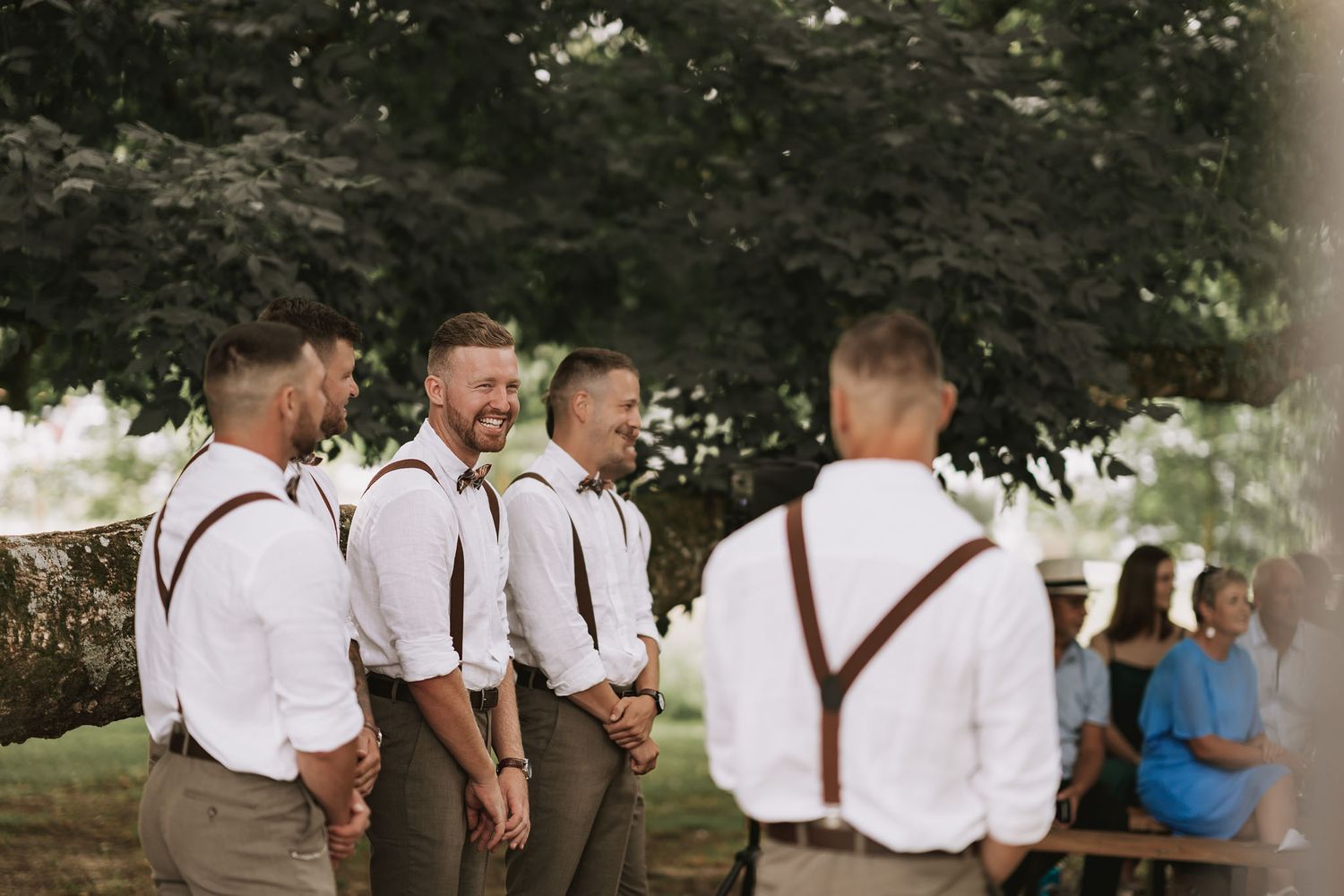 Groomsmen in brown pants and white shirts with suspenders share a laugh at an outdoor wedding ceremony.