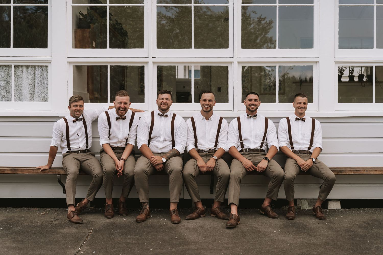 Six groomsmen in matching tan pants and suspenders sit casually on a railing outside a white building.