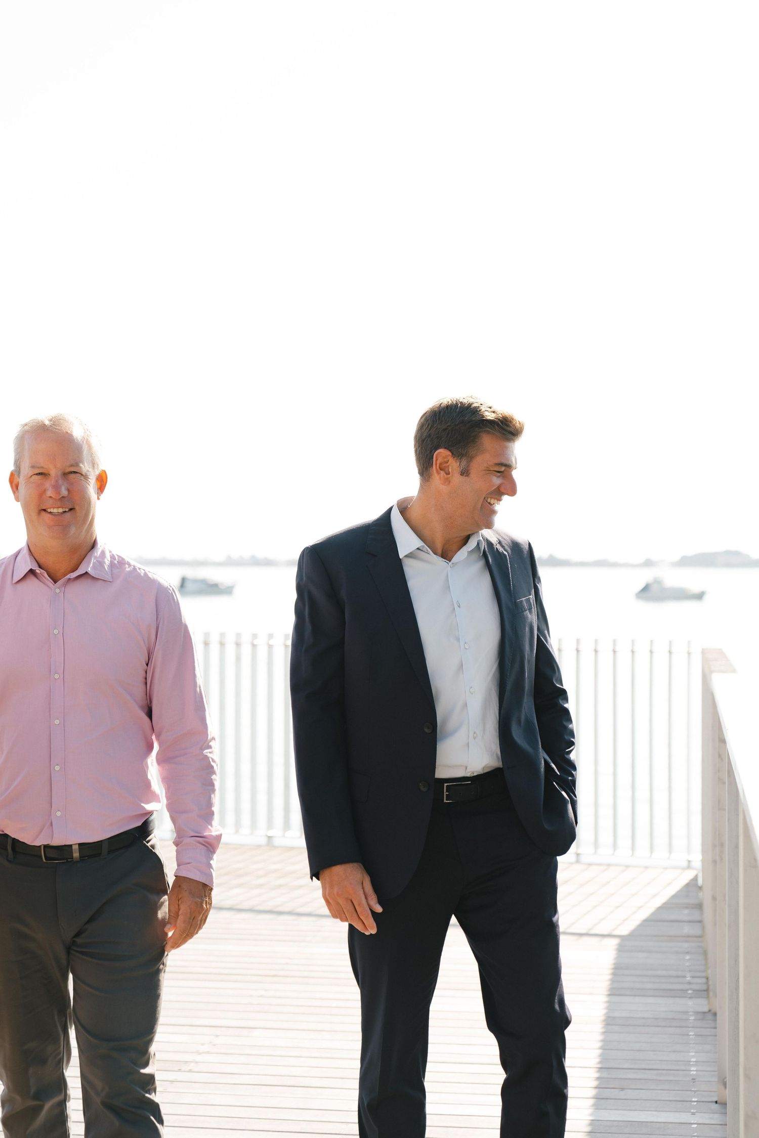 Two businessmen walking together along a sunny boardwalk, one wearing pink and one in navy suit.