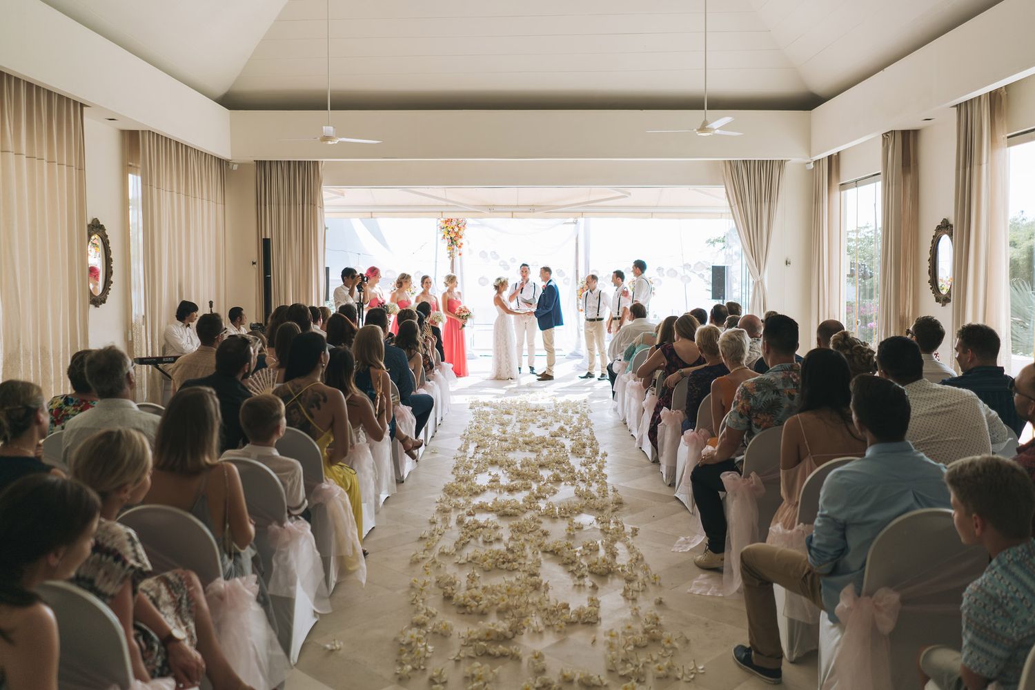 Indoor wedding ceremony with guests seated along a floral-lined aisle leading to oceanfront windows.
