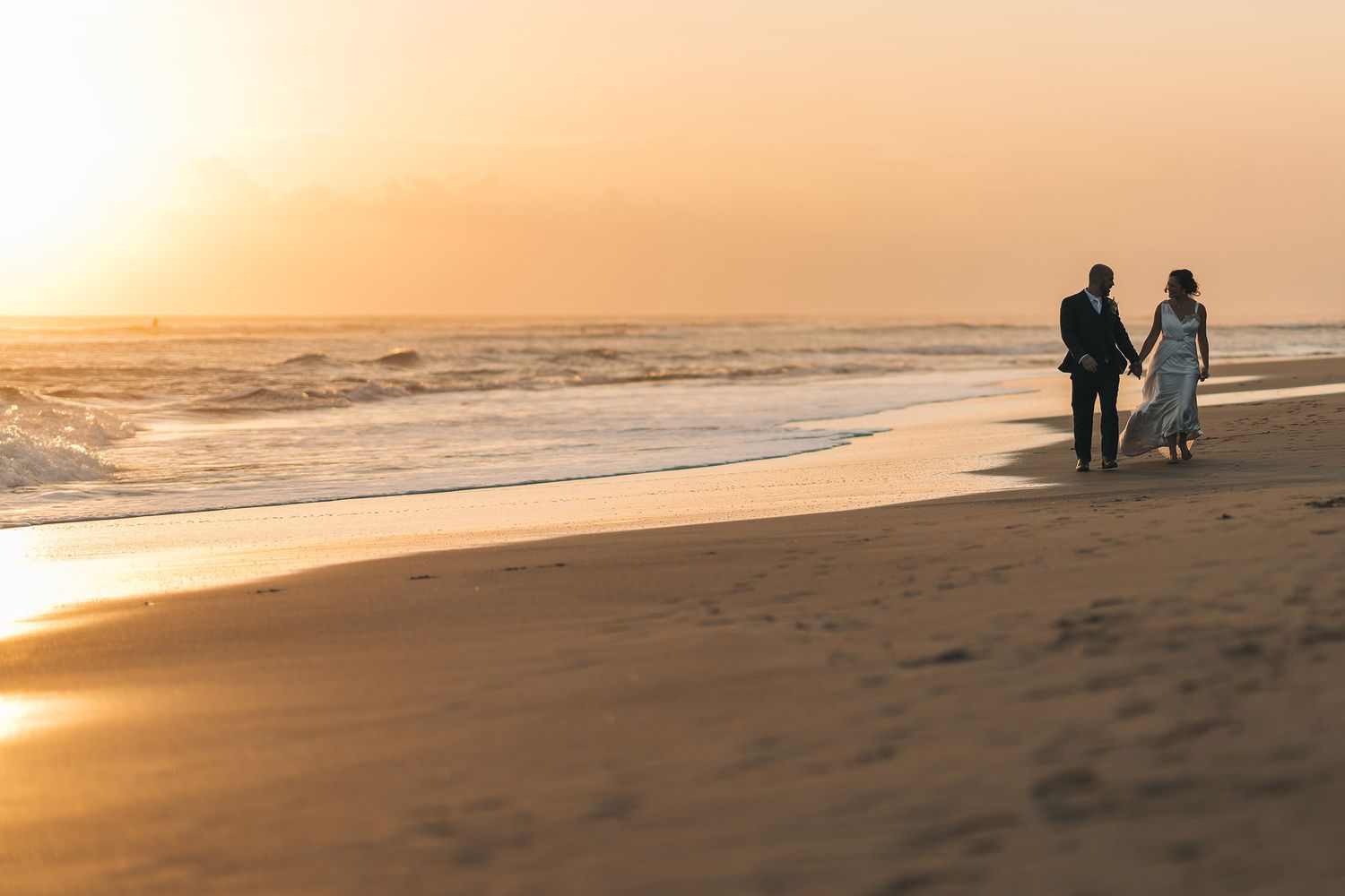 A silhouetted couple walks along a beach at sunset with golden light reflecting on the ocean waves.