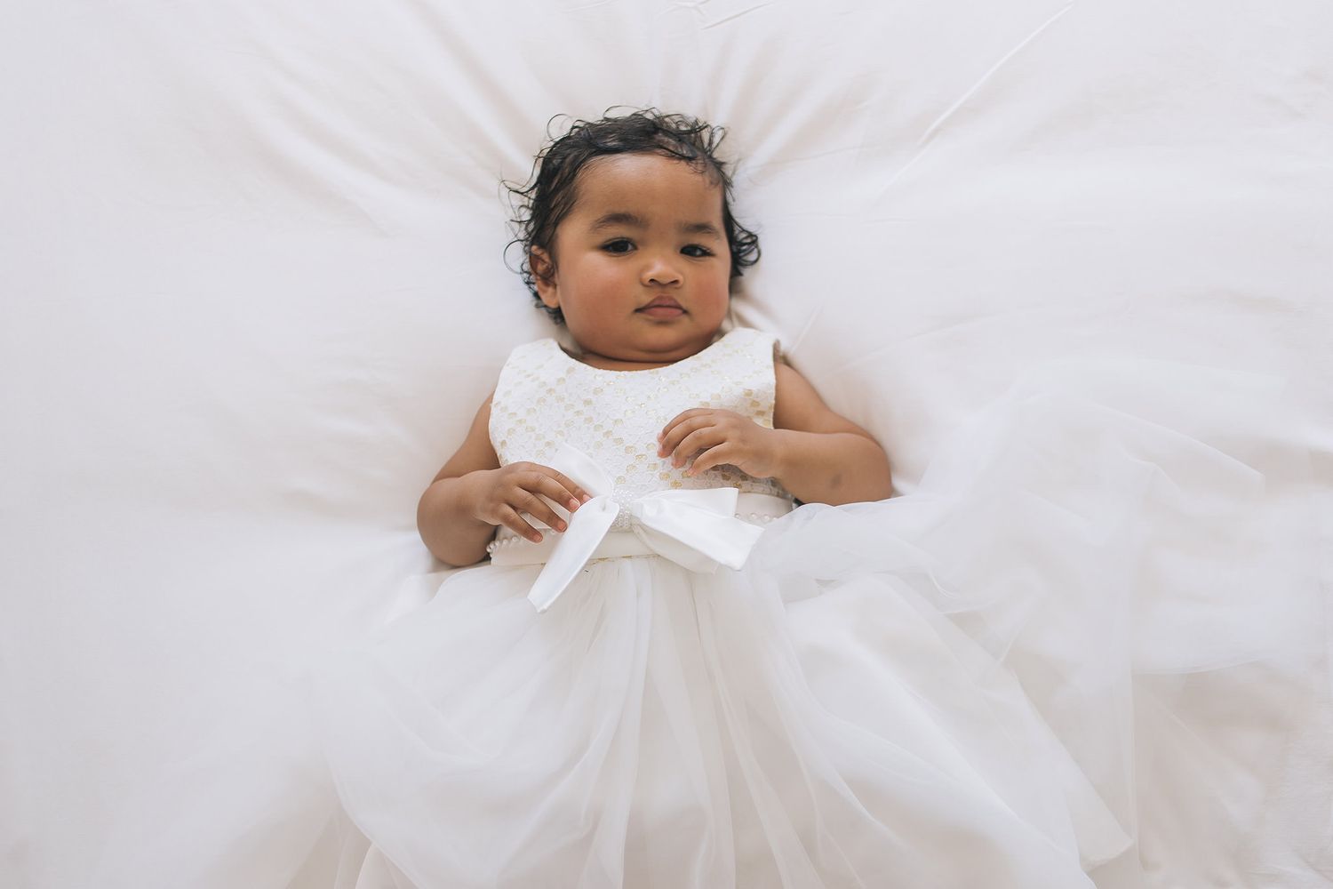 A baby wearing a white dress lies peacefully on white bedding, creating an angelic portrait.